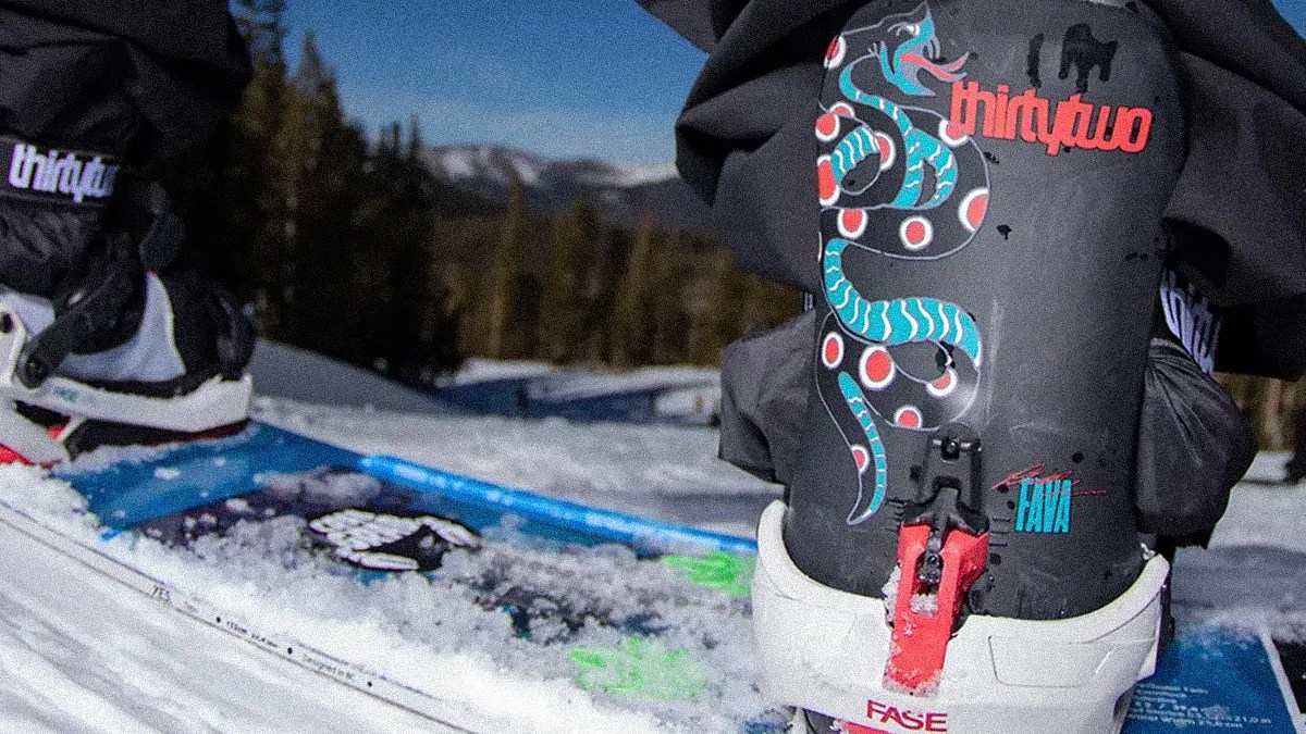 Close-up of a black ski boot with a colorful snake design and "thirtytwo" logo, attached to a blue ski on snow.