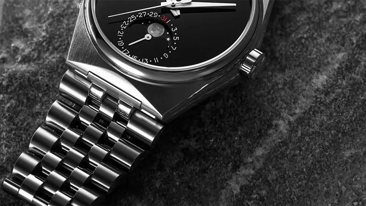Close-up of a stainless steel wristwatch with a black dial featuring a moon phase and date indicator on a dark textured surface.