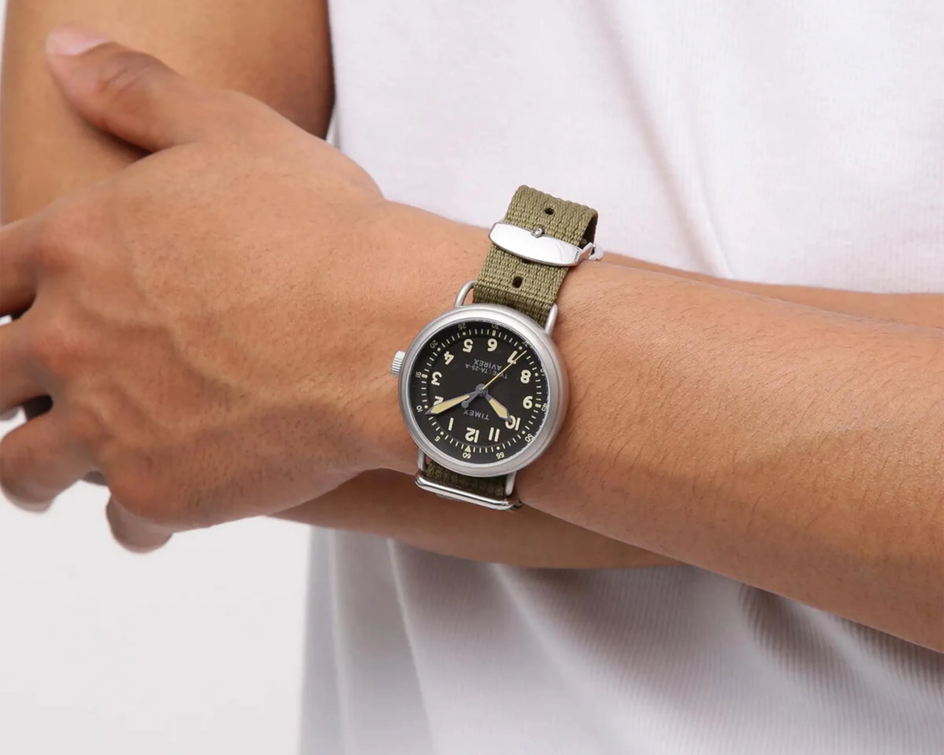 Silver Timex wristwatch with black dial and beige numerals on an olive green fabric strap worn on a wrist.