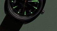 Close-up of a black wristwatch with luminous green hour markers and hands, black fabric strap, and silver crown.
