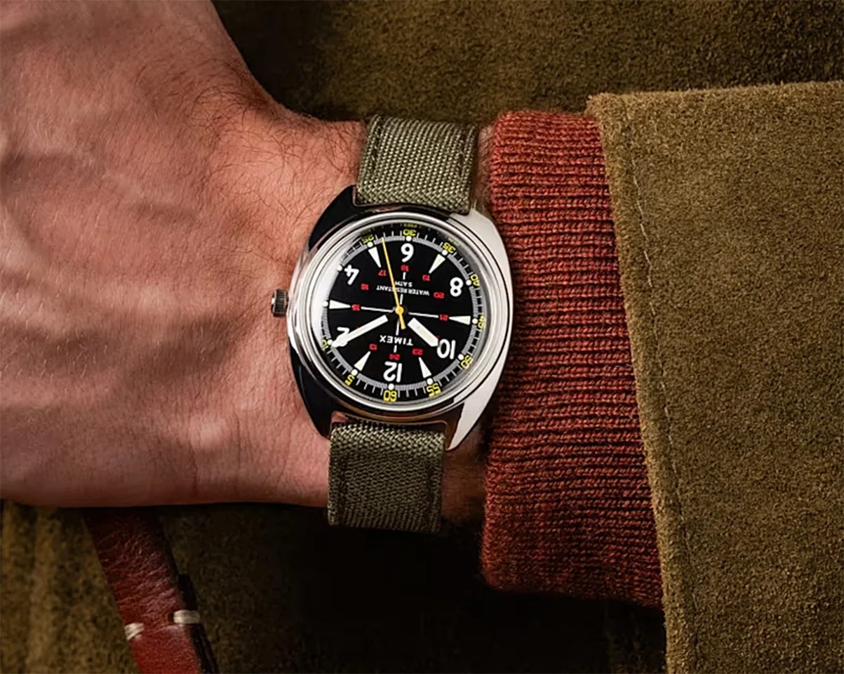 Timex wristwatch with black dial, white numerals, yellow second hand, and olive green fabric strap worn on a wrist.