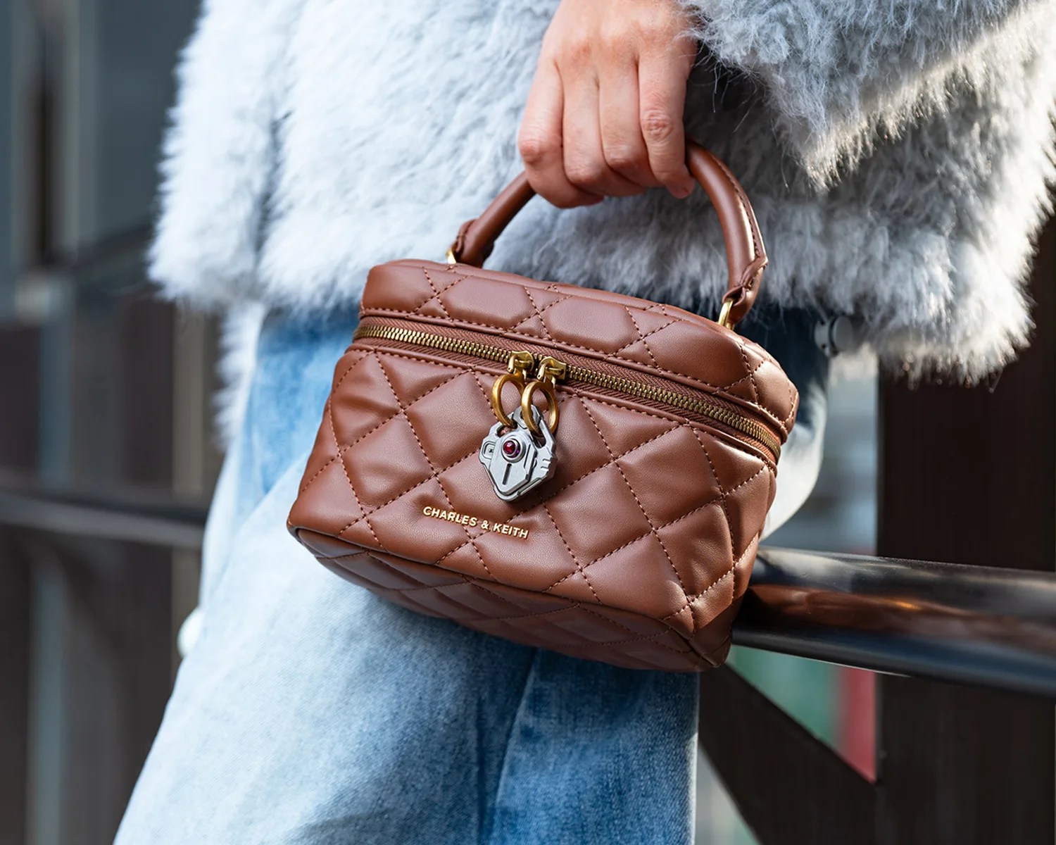 Brown quilted leather handbag with gold zipper and lock detail held by a person wearing a gray fuzzy sweater and blue jeans.