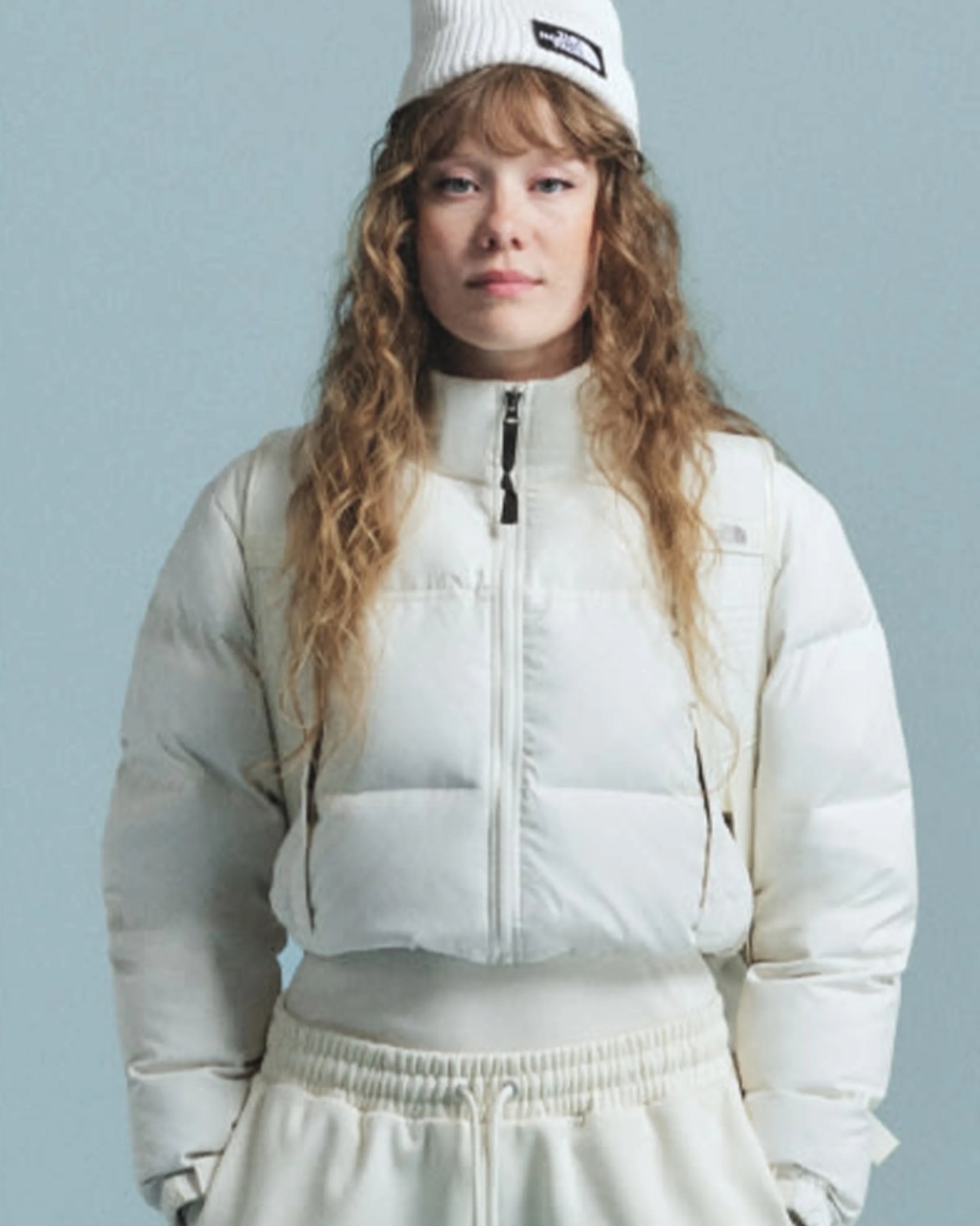 Woman wearing a white cropped puffer jacket, matching white beanie with a black logo, and white pants.