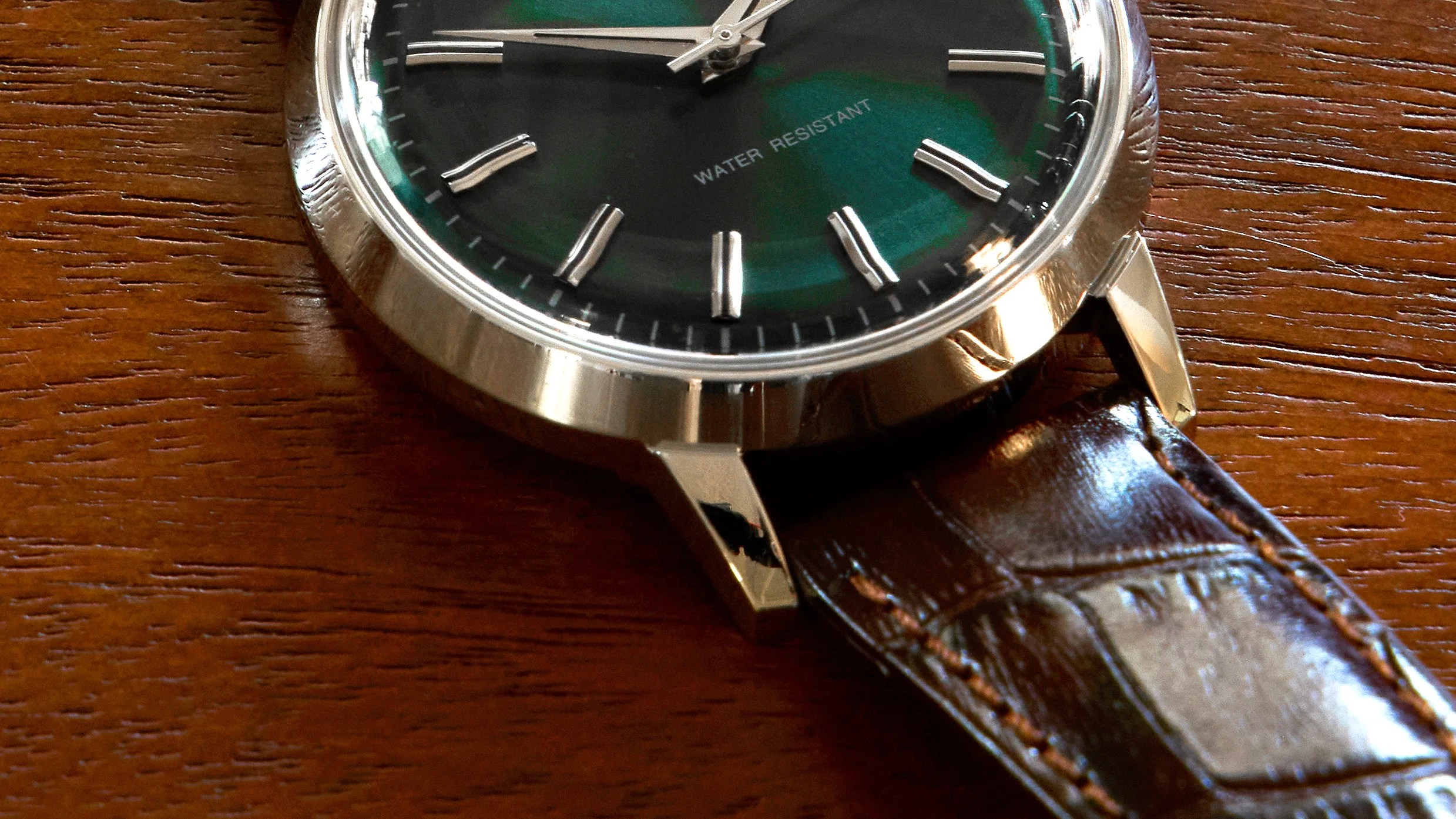 Close-up of a wristwatch with a silver case, green dial, and brown leather strap on a wooden surface.