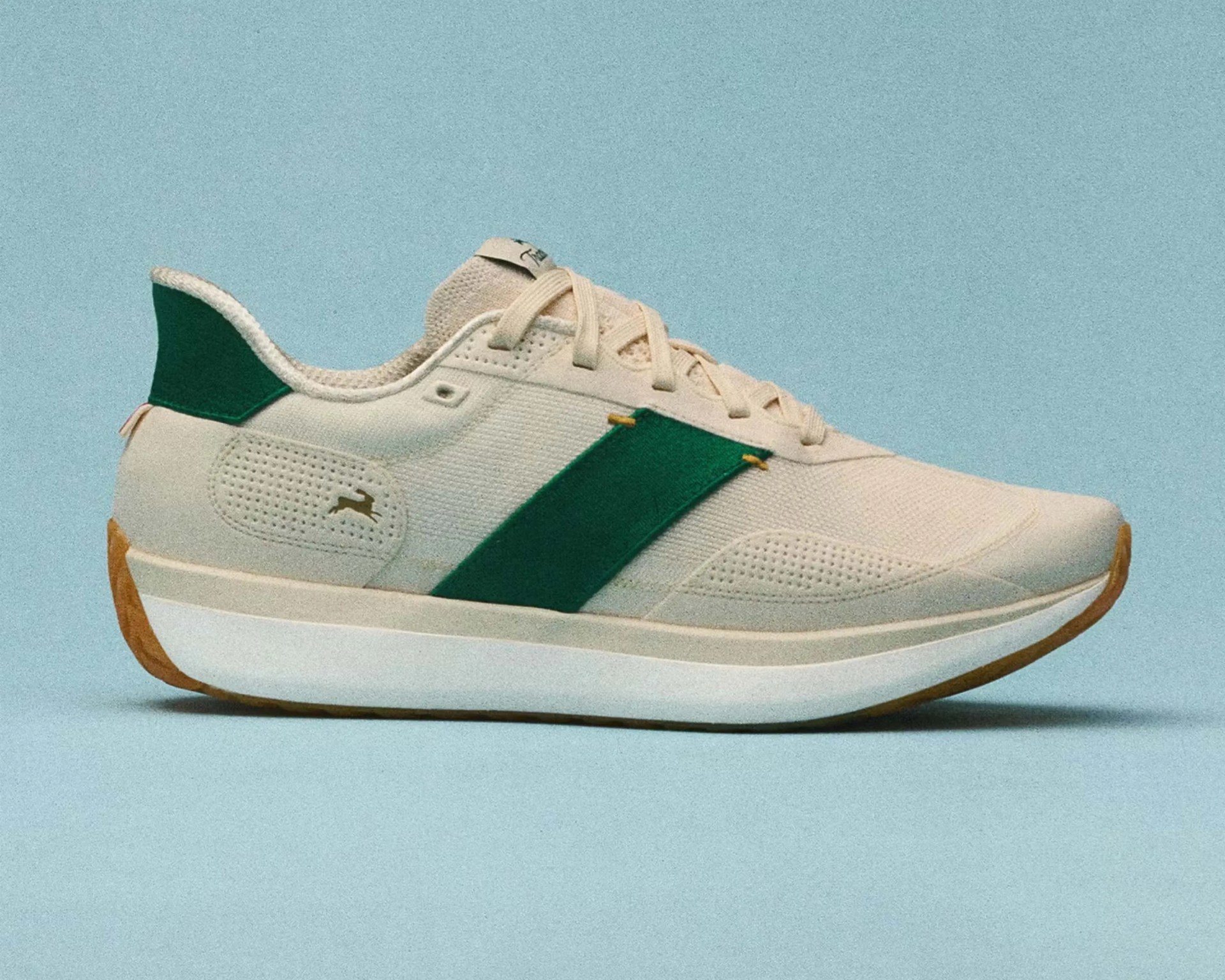 Beige sneaker with green side stripe, green heel tab, and white sole with gum outsole on a blue background.
