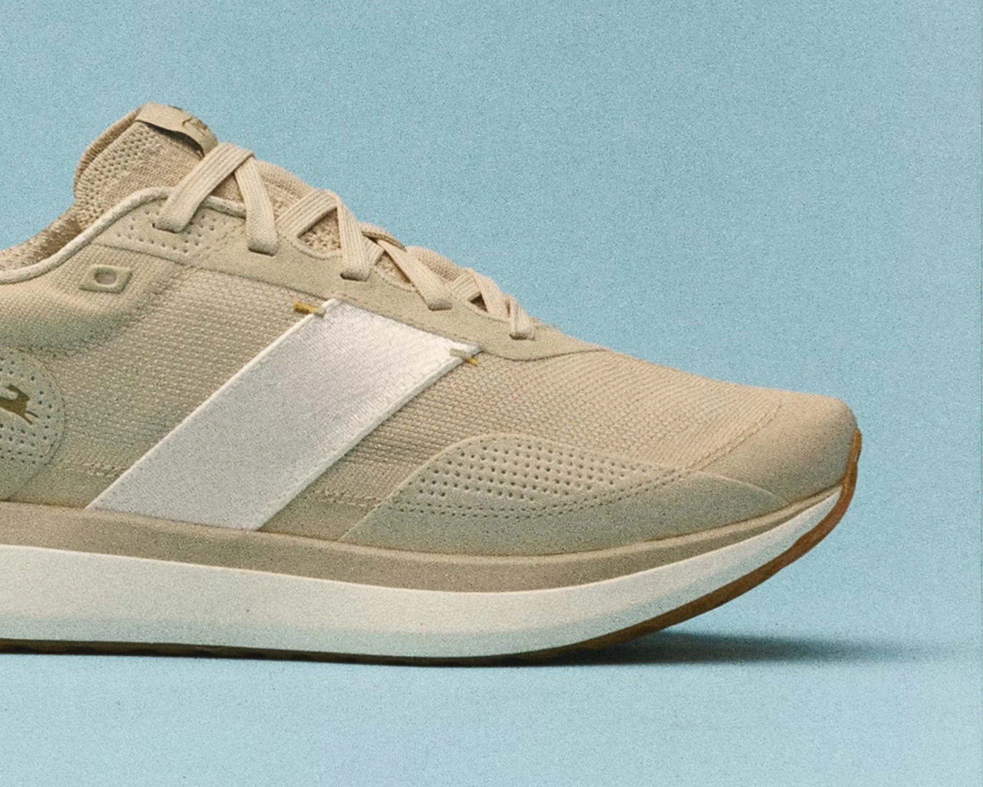 Beige athletic sneaker with white side stripe and textured sole on a light blue background.