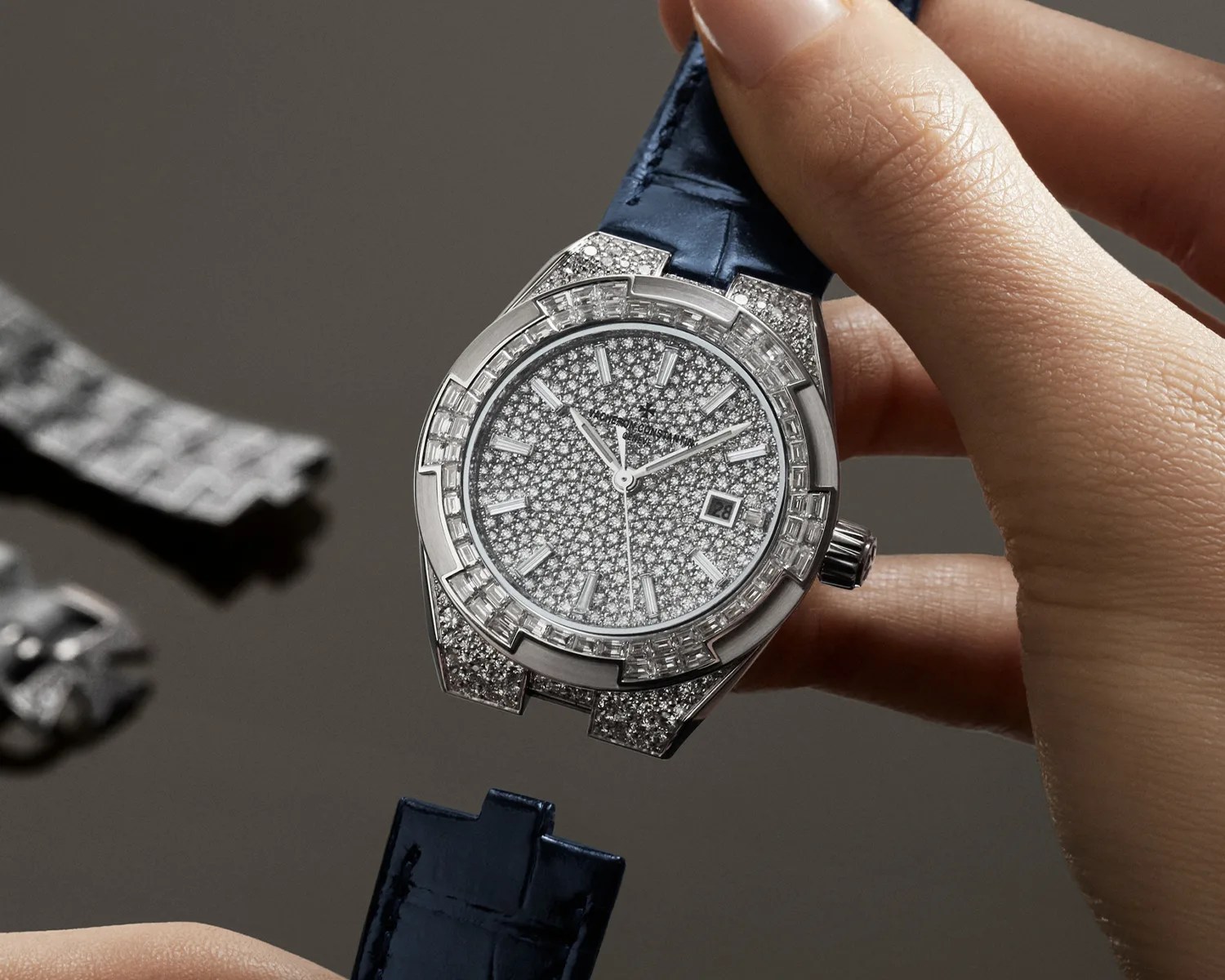 Hand holding a silver luxury watch with a diamond-encrusted face and bezel, and a detached dark blue leather strap.