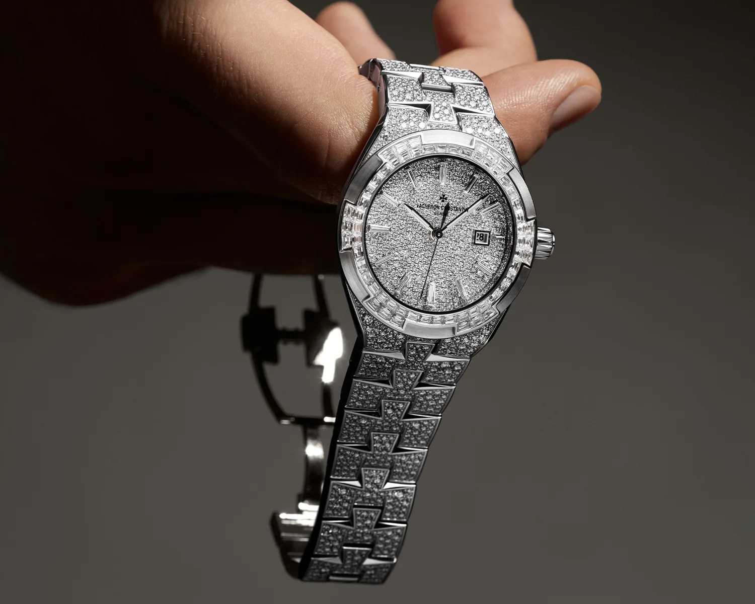 Silver luxury wristwatch encrusted with diamonds held in a hand against a dark background.