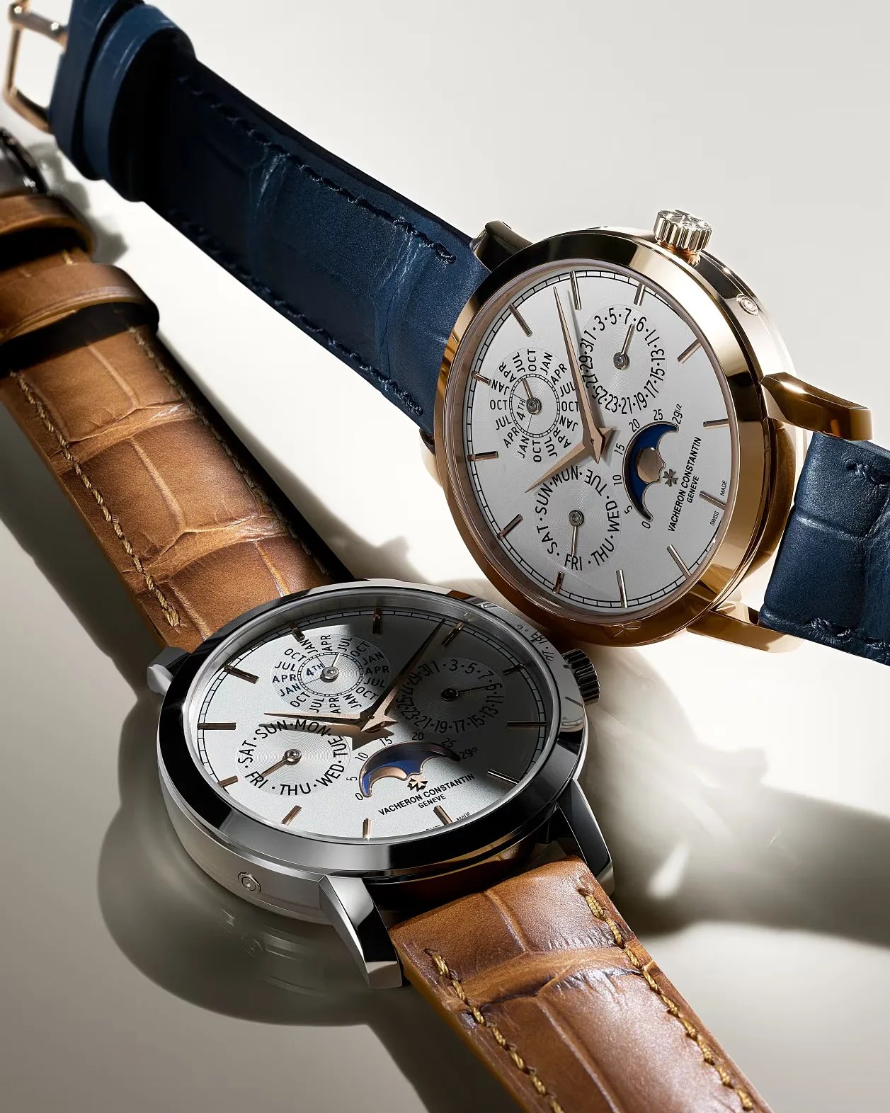 Two luxury wristwatches with moon phase and calendar dials, one with a blue leather strap and gold case, the other with a brown leather strap and silver case.
