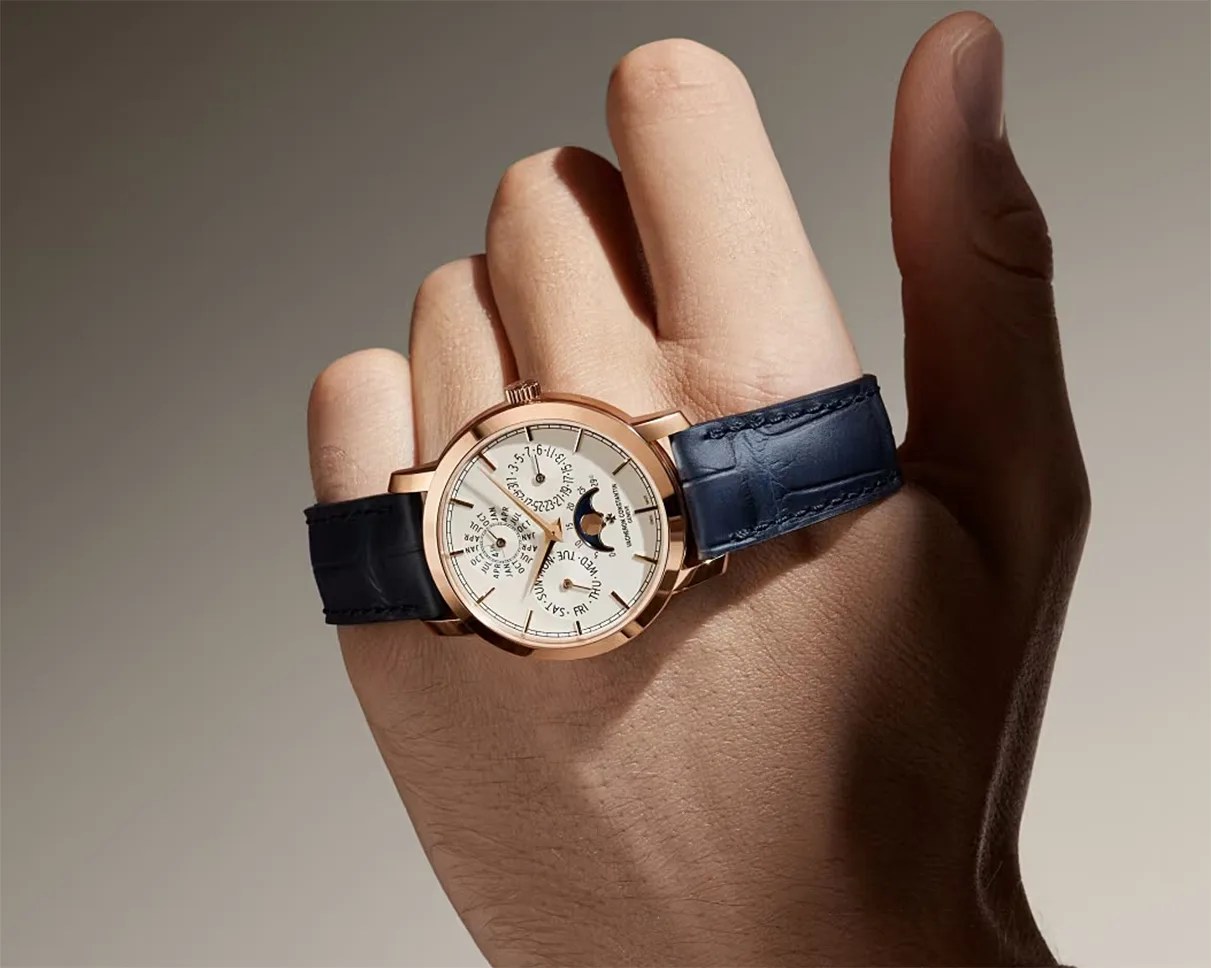 Rose gold wristwatch with white dial, moon phase, and blue leather strap held in a hand.