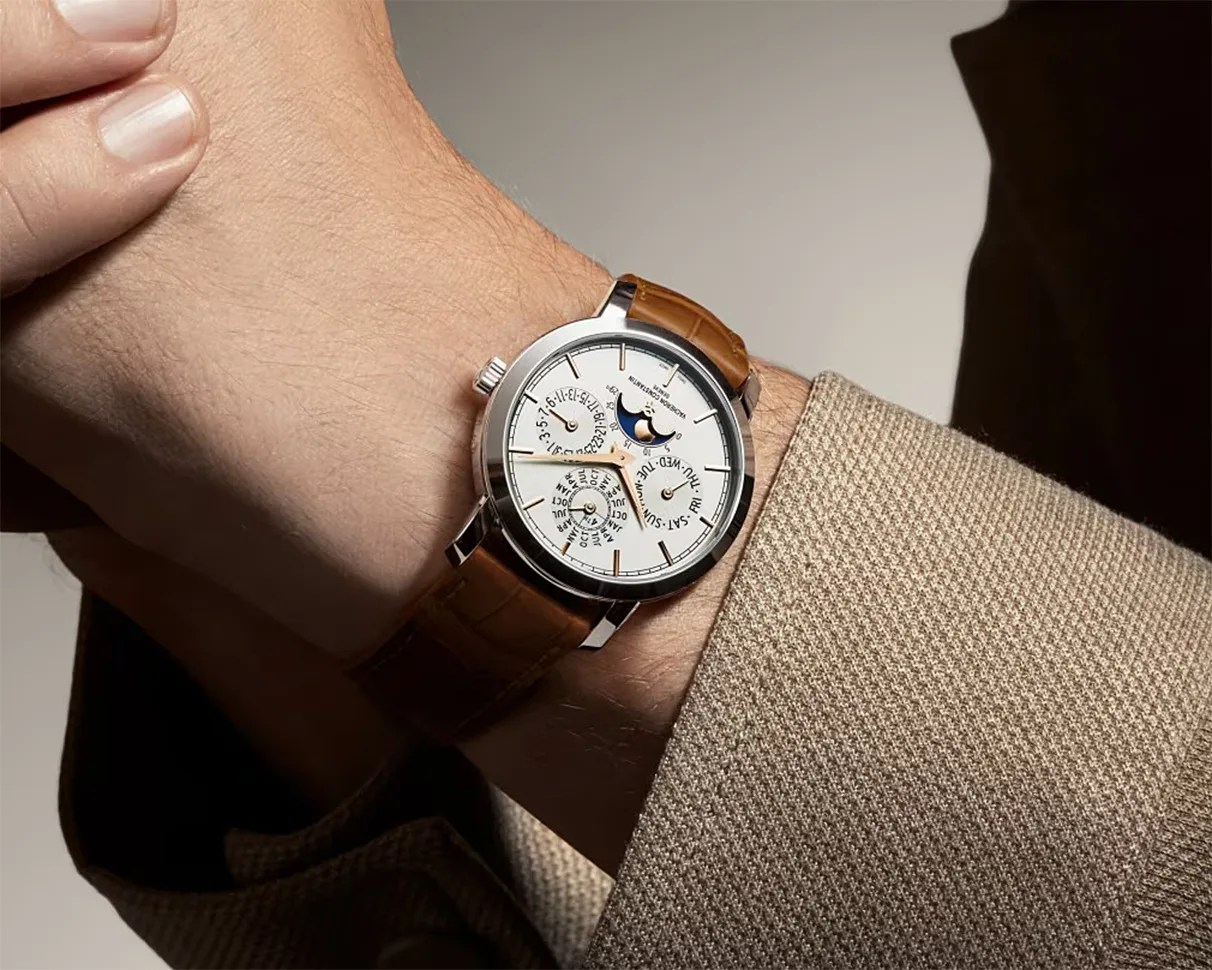 Silver wristwatch with white dial, moon phase indicator, and brown leather strap worn on a wrist with a beige textured sleeve.