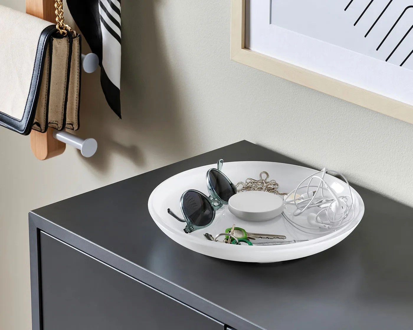 White ceramic dish holding sunglasses, keys, earbuds, and a small round device on a dark gray cabinet.