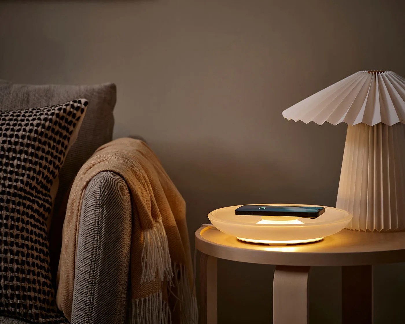 Round frosted glass wireless charger with warm light on a wooden side table next to a beige sofa with a blanket.