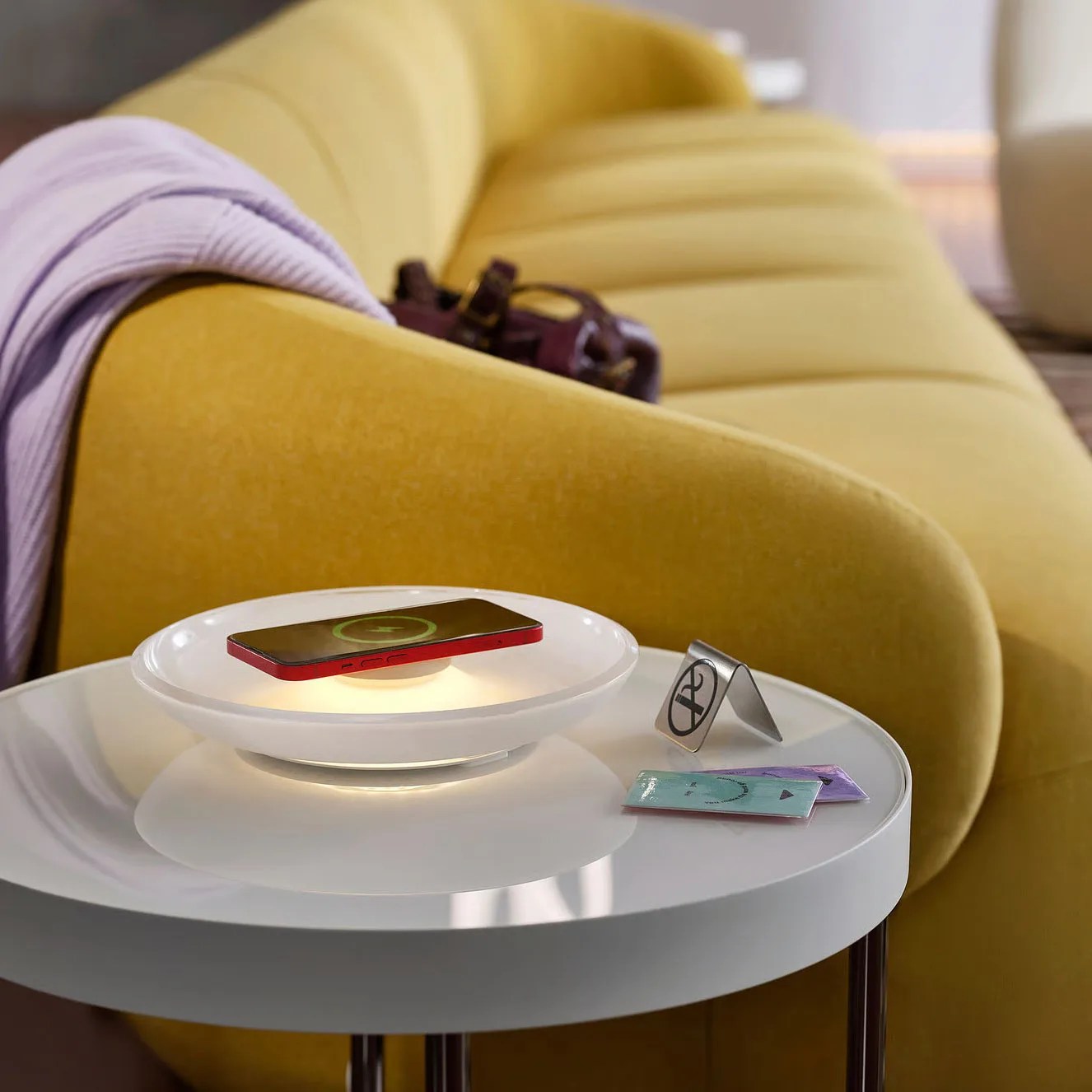 White round side table with a wireless charging pad holding a red phone, next to a no-smoking sign and two small packets, in front of a yellow sofa with a purple blanket.