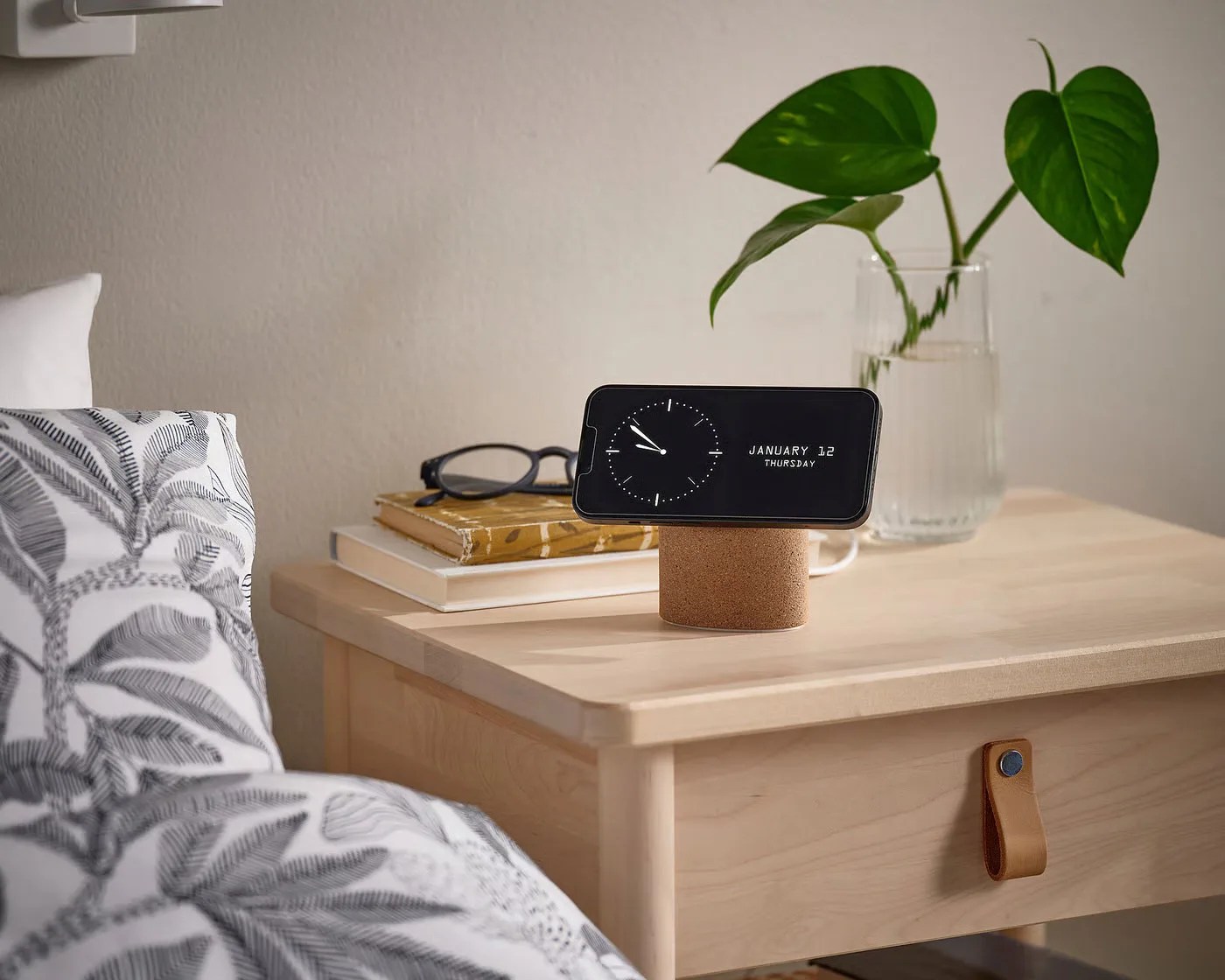Smartphone displaying clock and date on a cork stand on a light wood bedside table with books, glasses, and a glass vase with green leaves.