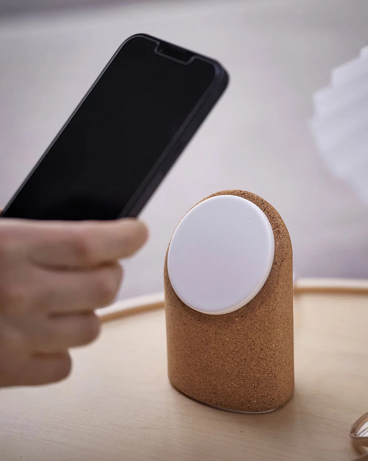 Brown cork wireless charger with a white circular charging pad on a wooden surface, phone held nearby.