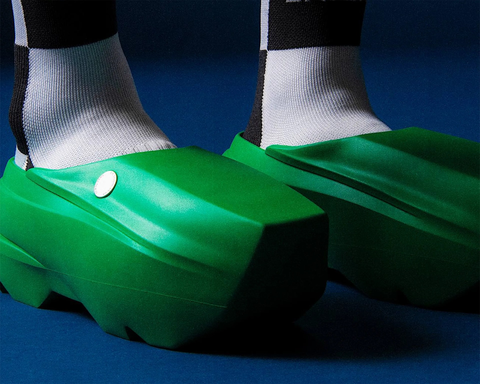 Chunky green slip-on shoes with a geometric design worn with black and white socks on a blue surface.