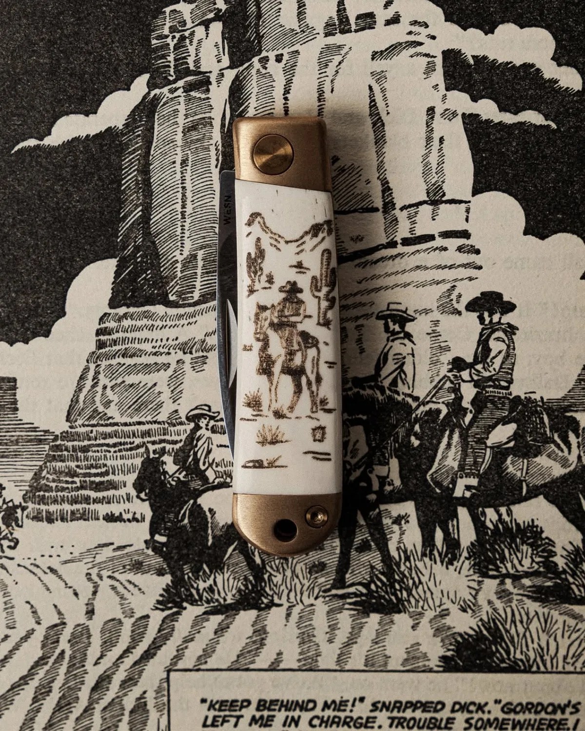 Folding pocket knife with a white handle featuring a cowboy on horseback illustration, set against a black-and-white Western comic background.
