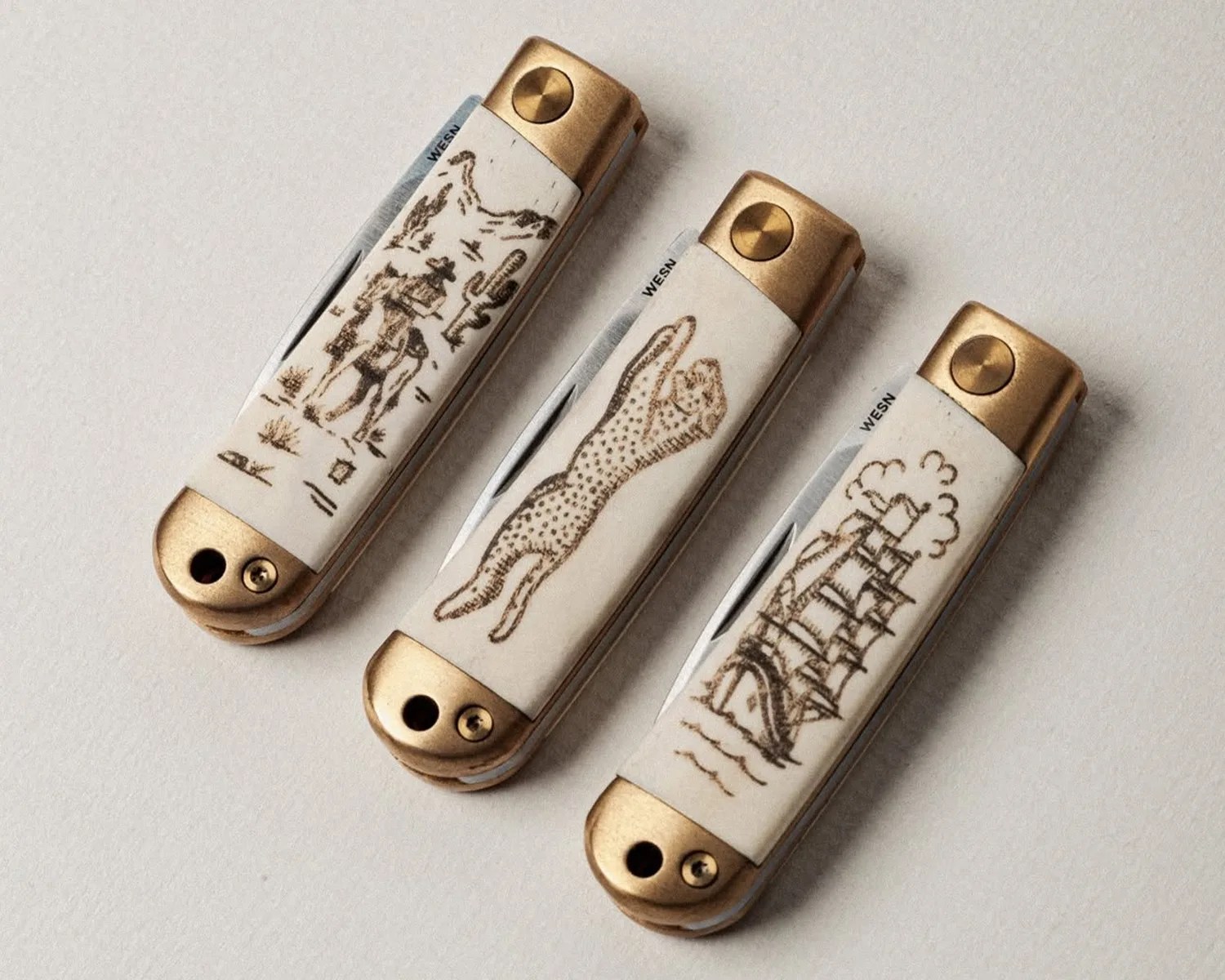Three folding knives with brass bolsters and white handles featuring engraved designs of a cowboy on horseback, a leaping leopard, and a sailing ship.