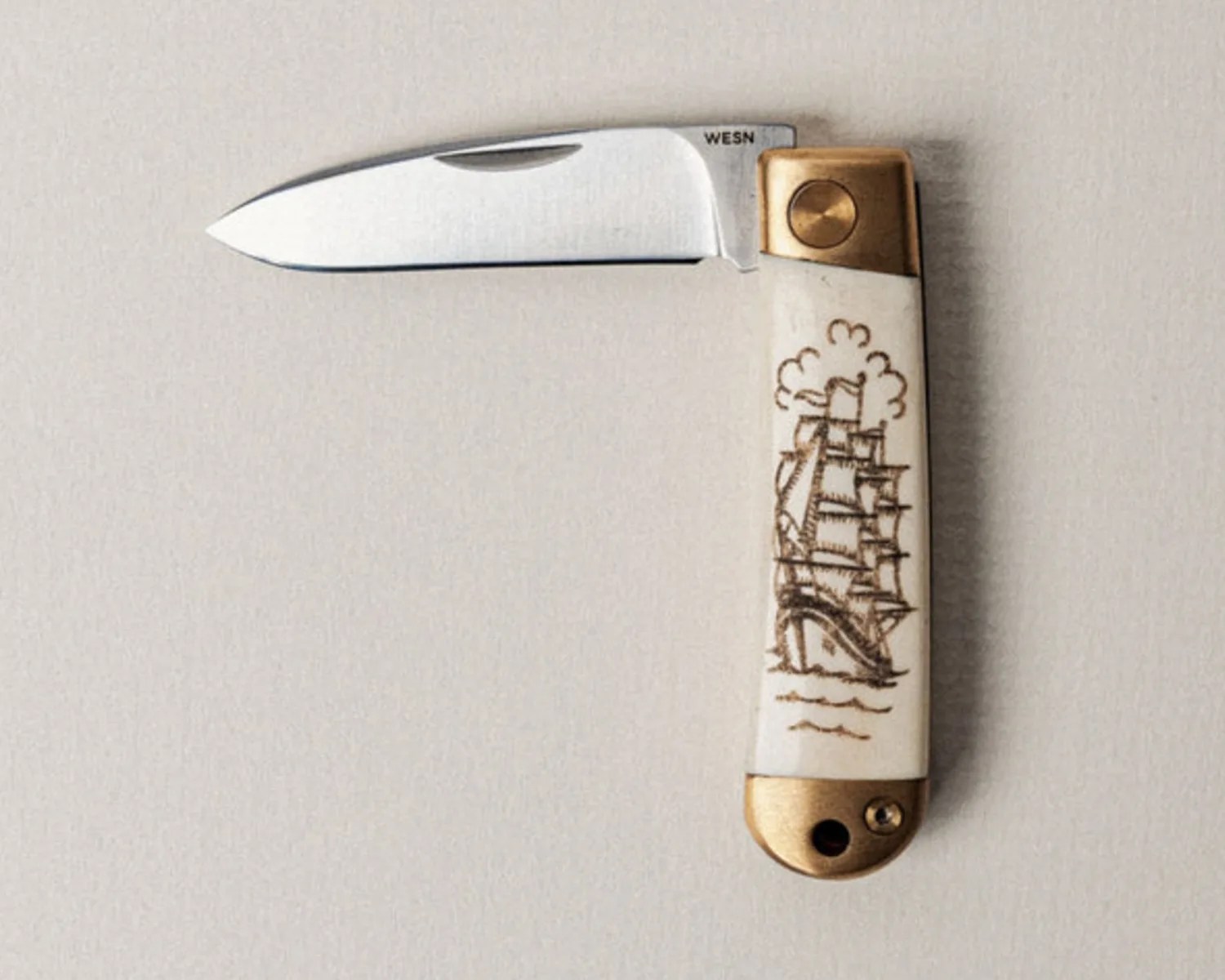 Folding pocket knife with a silver blade and a white handle featuring a brown ship engraving and brass accents.