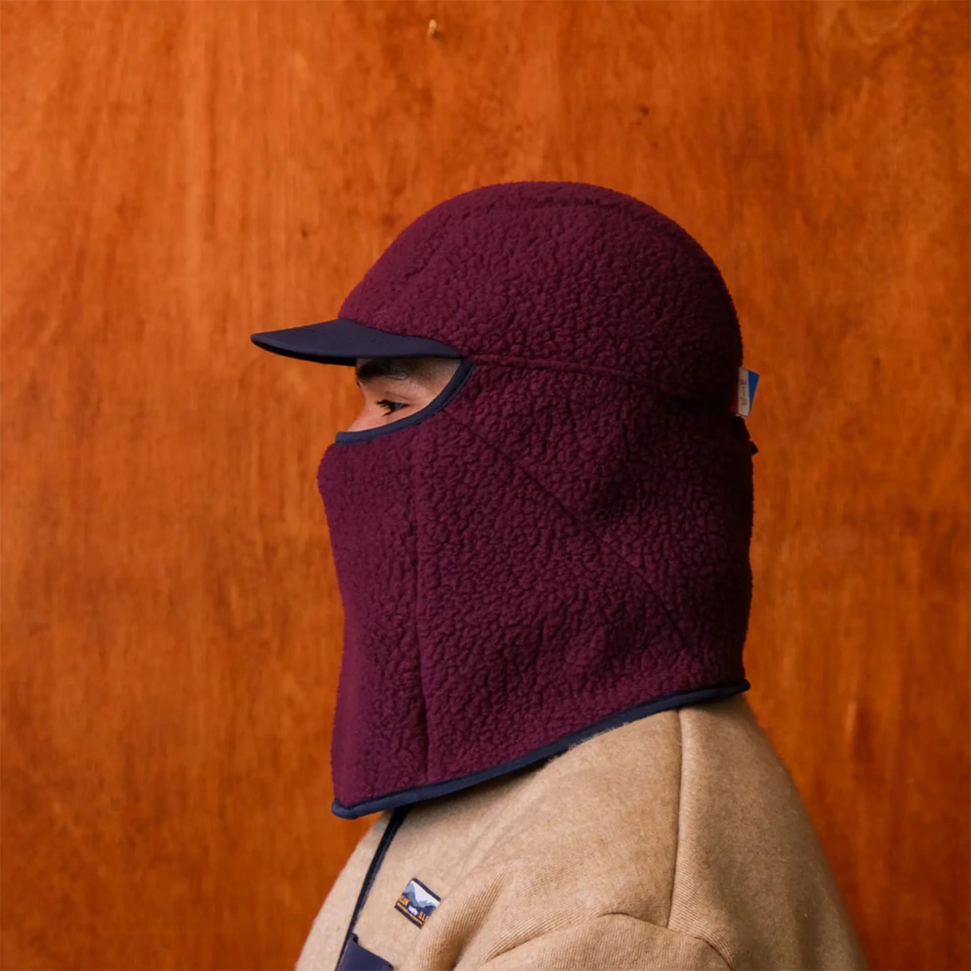 Person wearing a maroon fleece balaclava with a navy blue brim and trim against a wooden background.