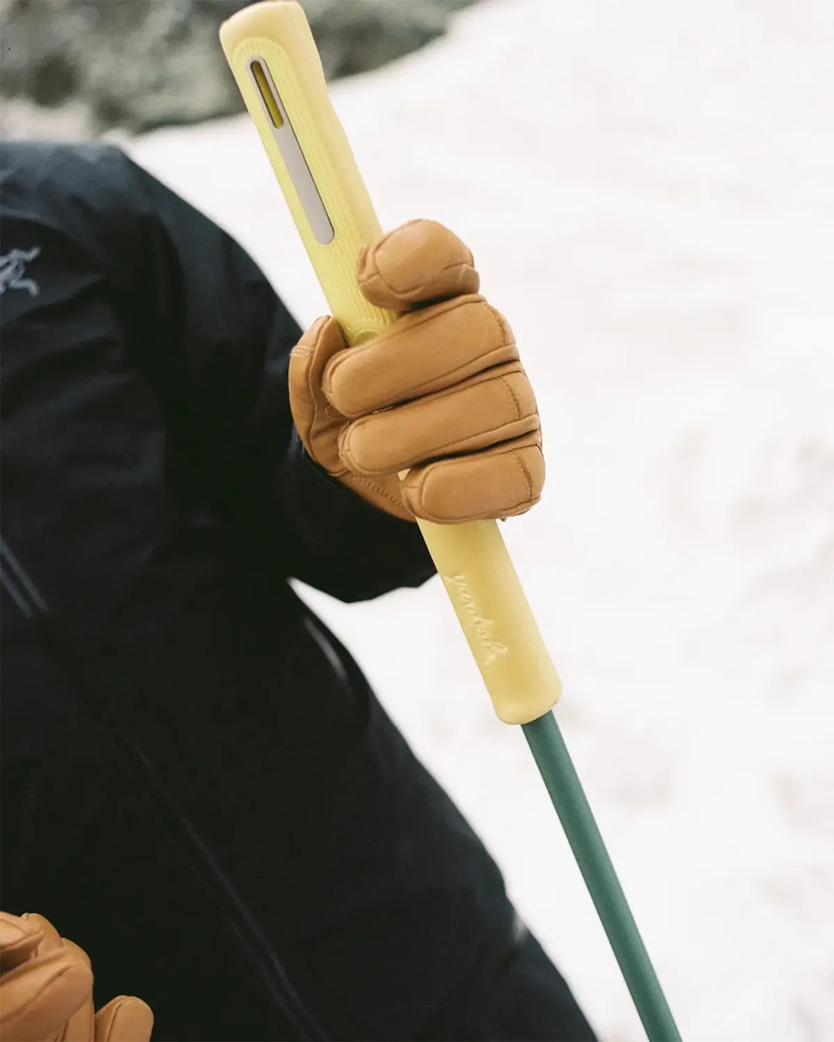 Person wearing tan leather gloves holding a yellow ski pole handle with a green shaft.