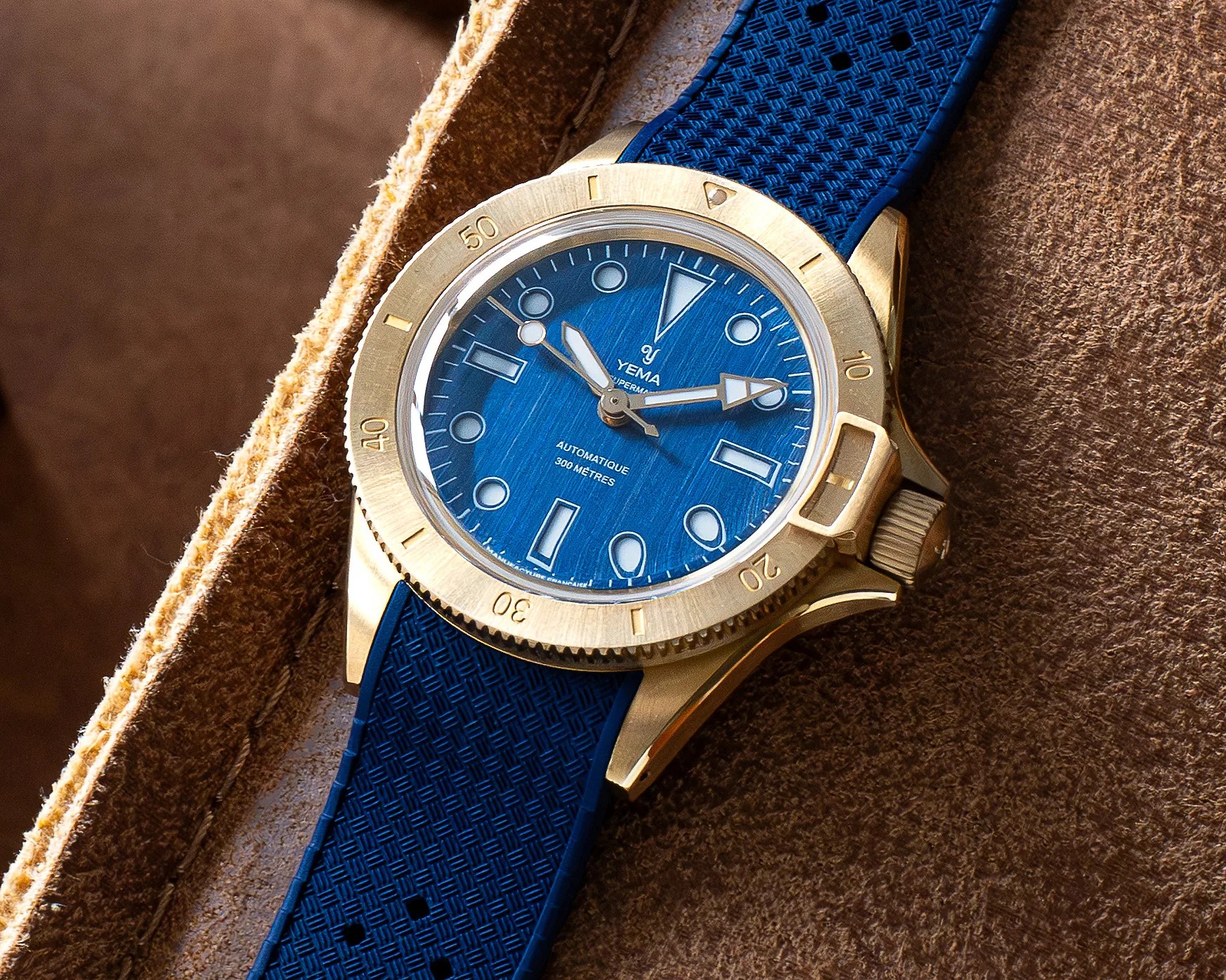 Gold-tone Yema dive watch with blue textured dial and matching blue rubber strap on brown leather surface.