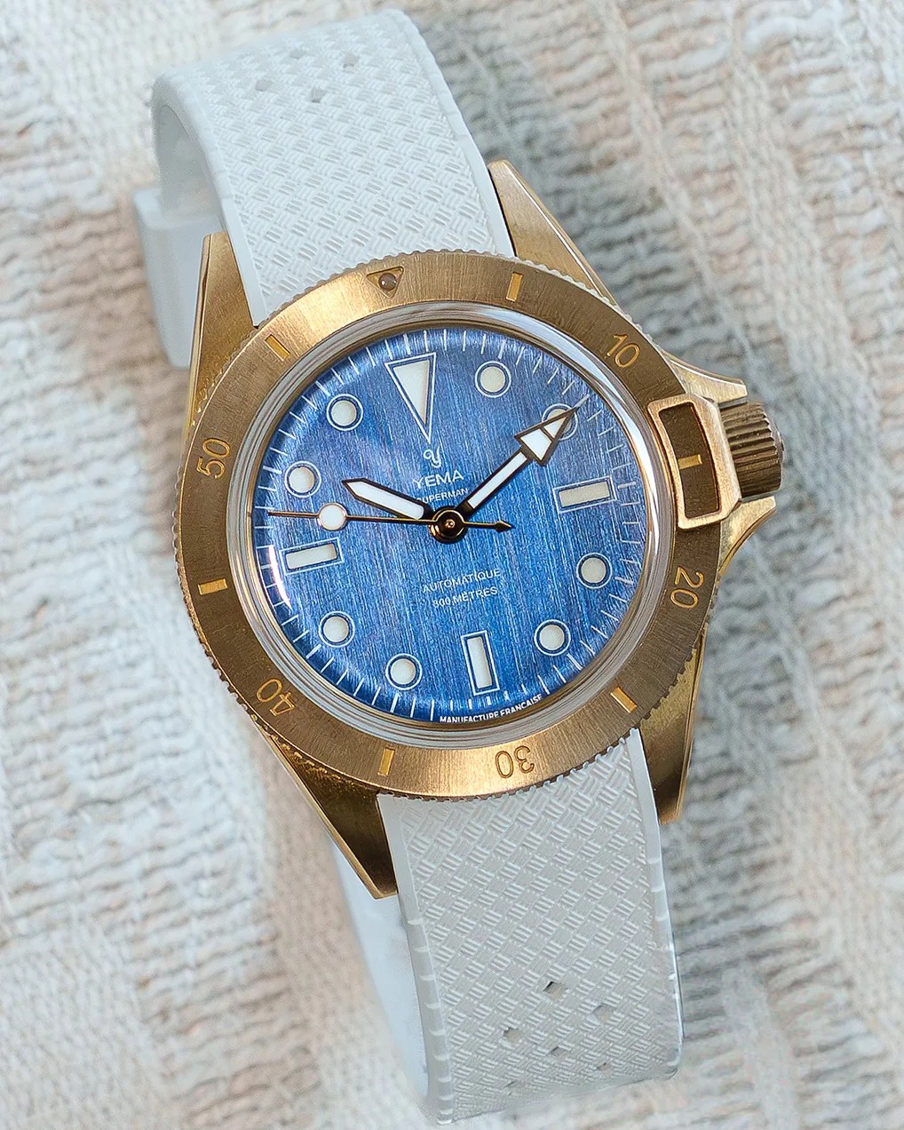 Diver-style wristwatch with a bronze case, blue textured dial, gold bezel, and white textured rubber strap.