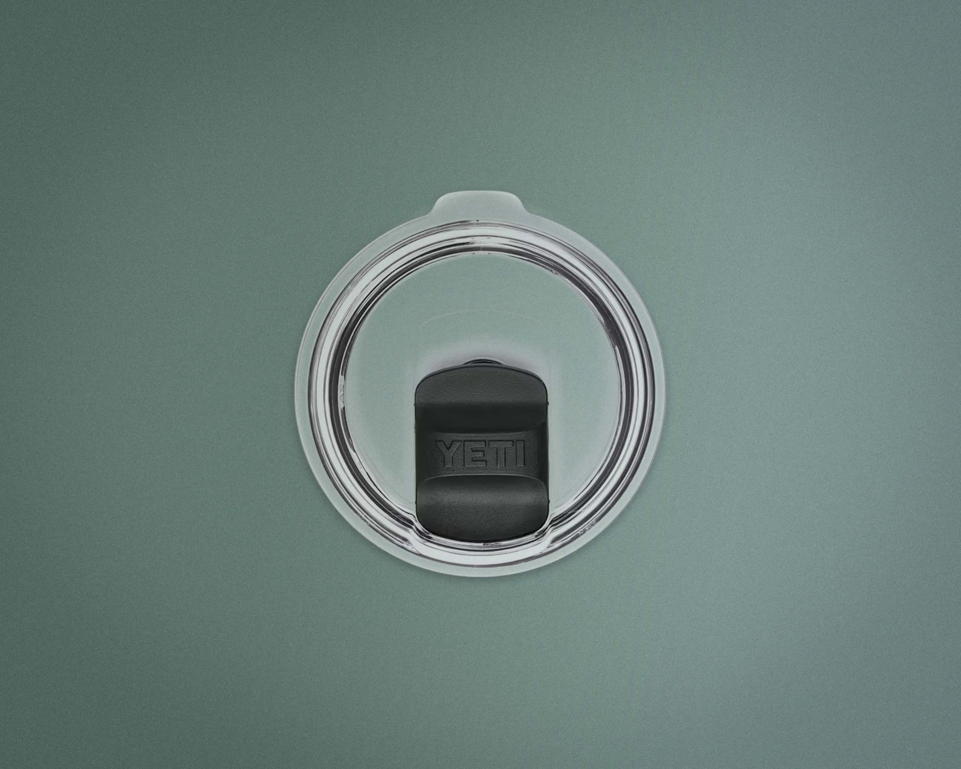 Clear circular YETI tumbler lid with black sliding closure on a green background.