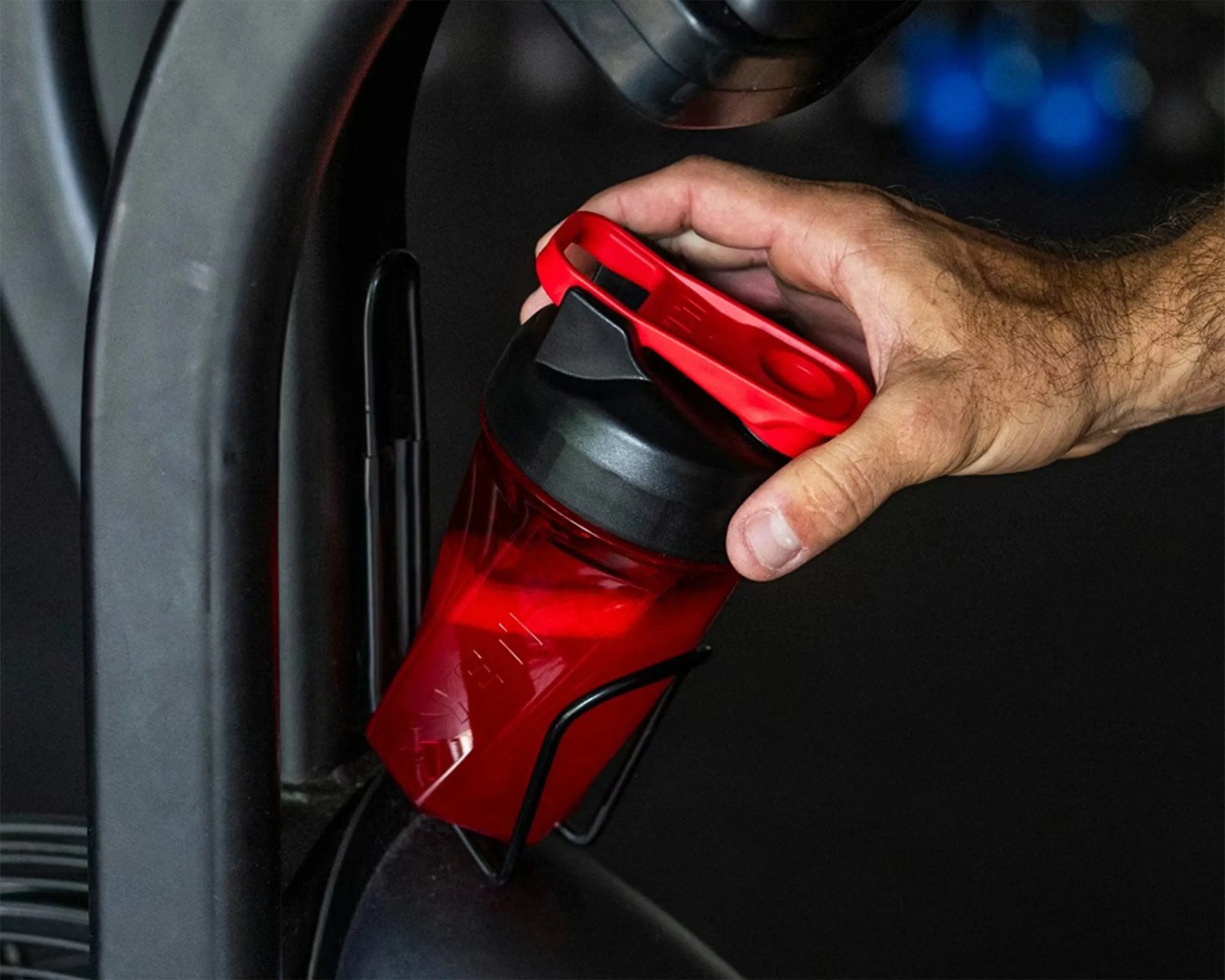 Hand placing a red and black shaker bottle into a black metal holder on gym equipment.