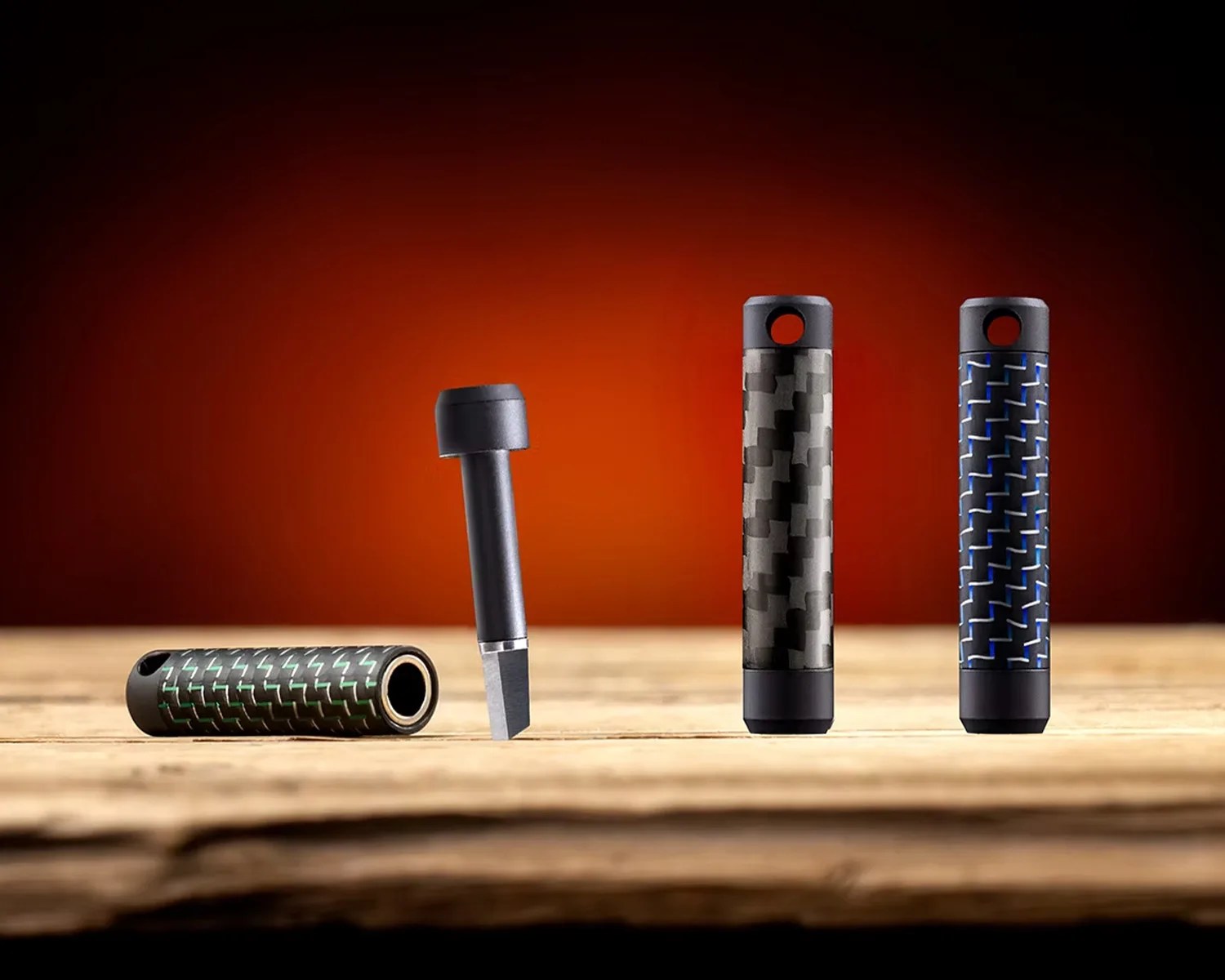 Three cylindrical carbon fiber keychain holders in green, black, and blue patterns, with one holder open showing a small metal tool inside.