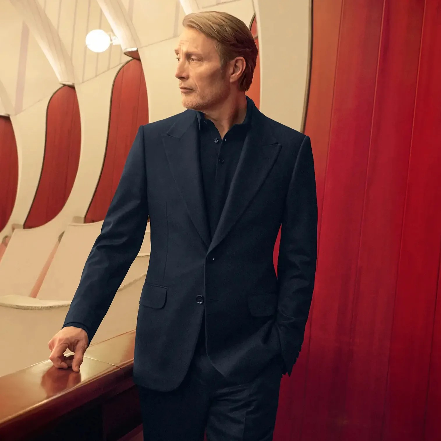 Man wearing a dark navy suit with a matching shirt, standing indoors against a red and beige wall.