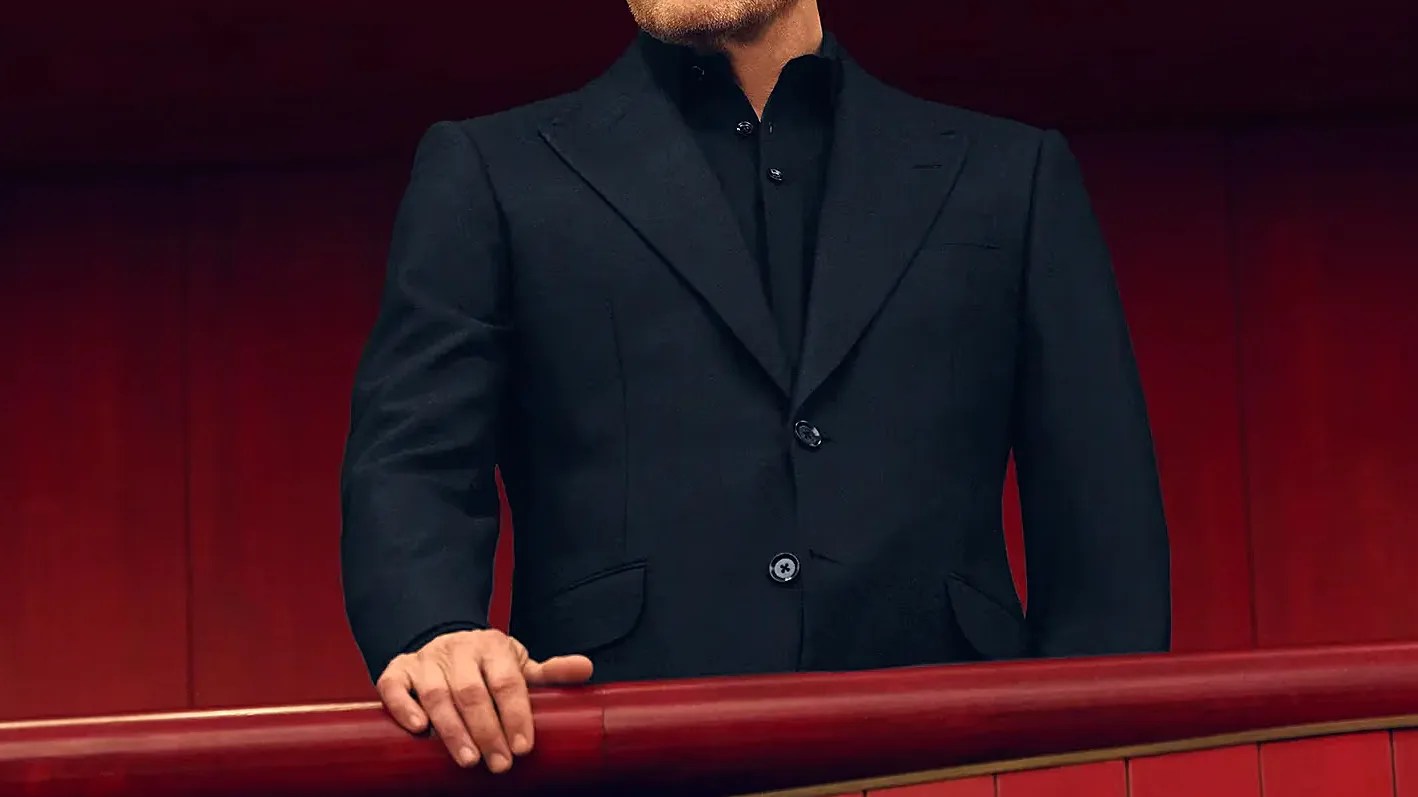 Man wearing a black suit jacket and black shirt standing behind a red railing.