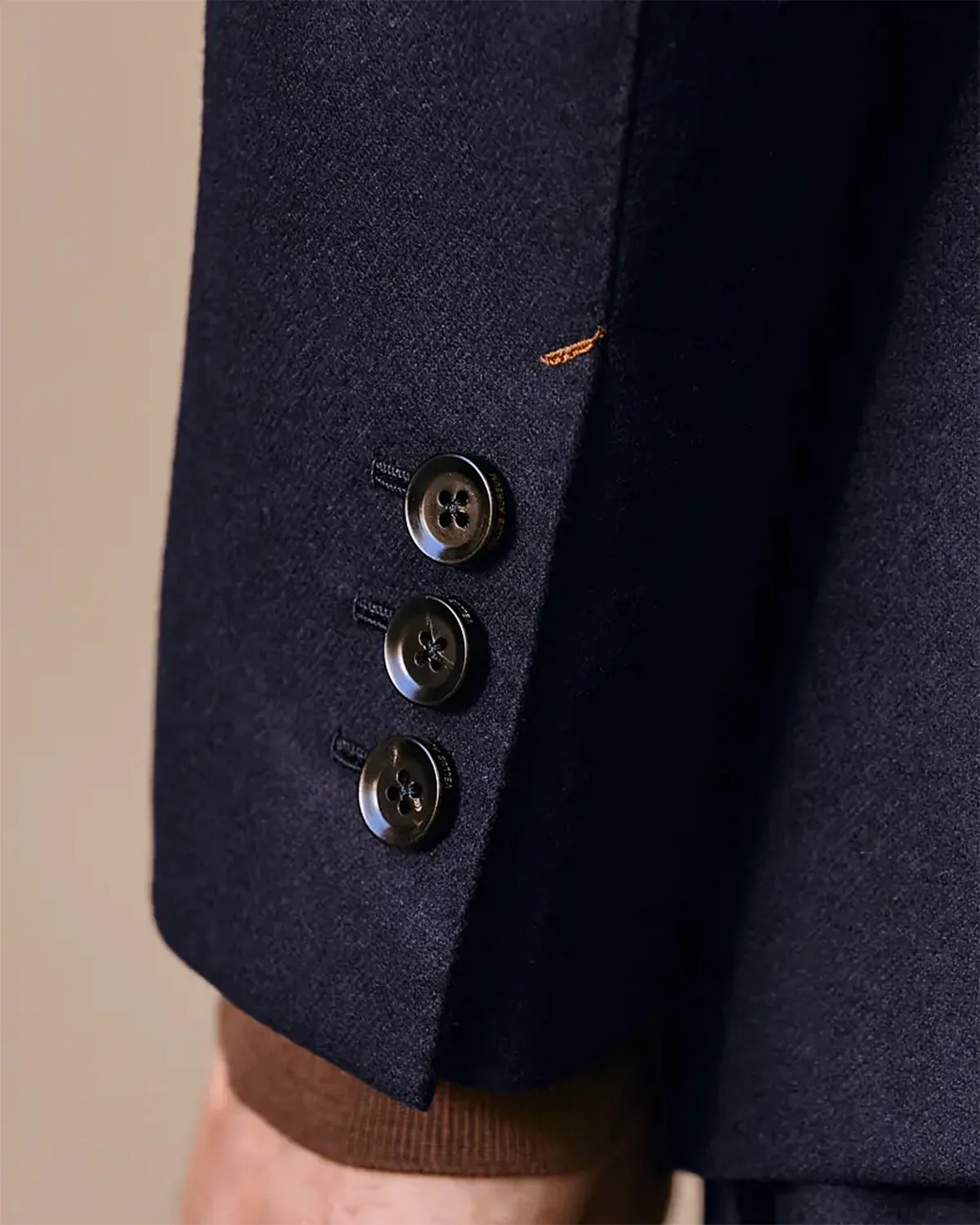 Close-up of a navy blue blazer sleeve with three black buttons and a small orange stitch detail.