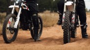 Two dirt bikes with knobby tires on a sandy trail, riders wearing black gear and boots.