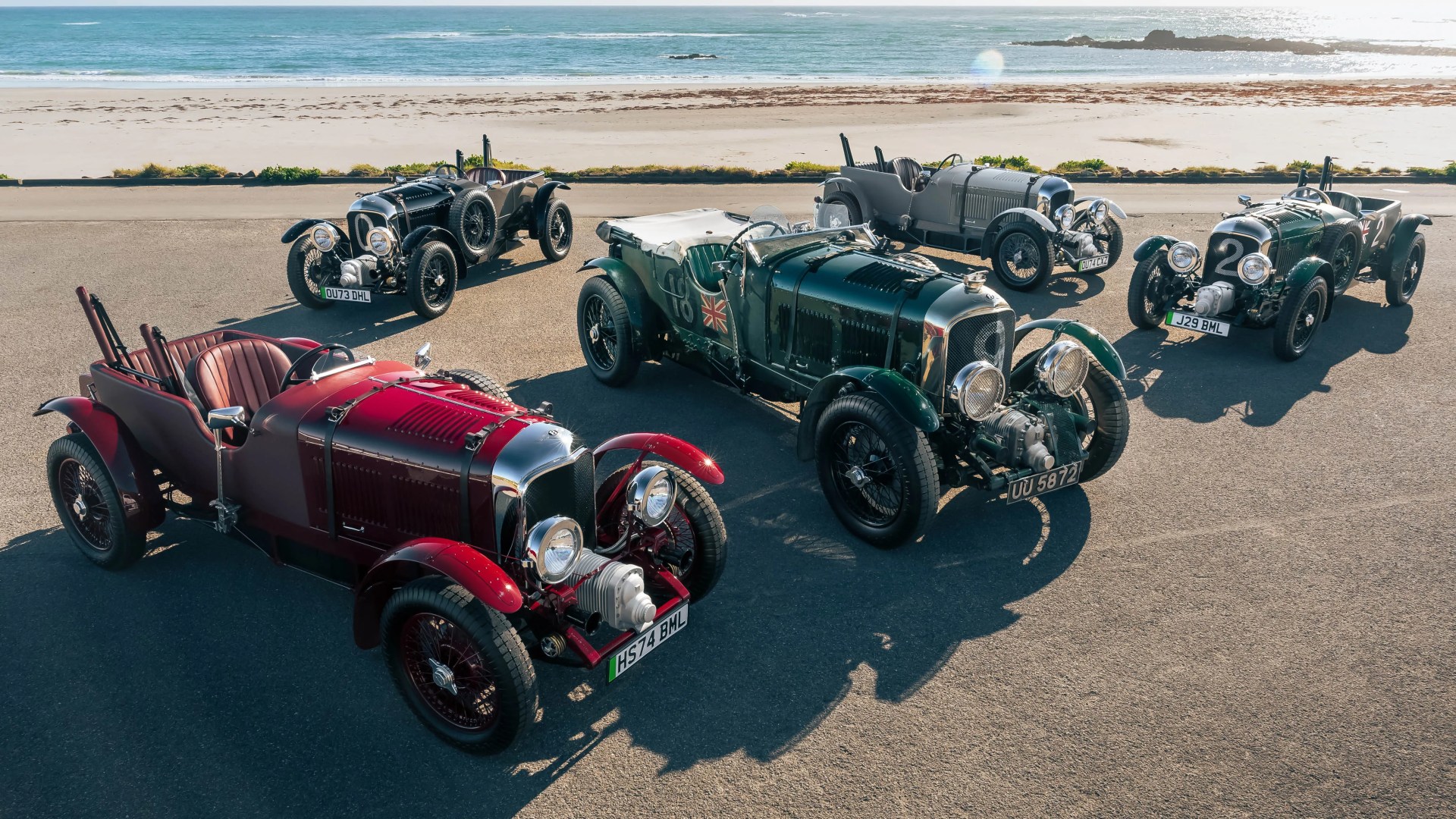 Five vintage open-top racing cars in various colors parked on a paved area near a beach.