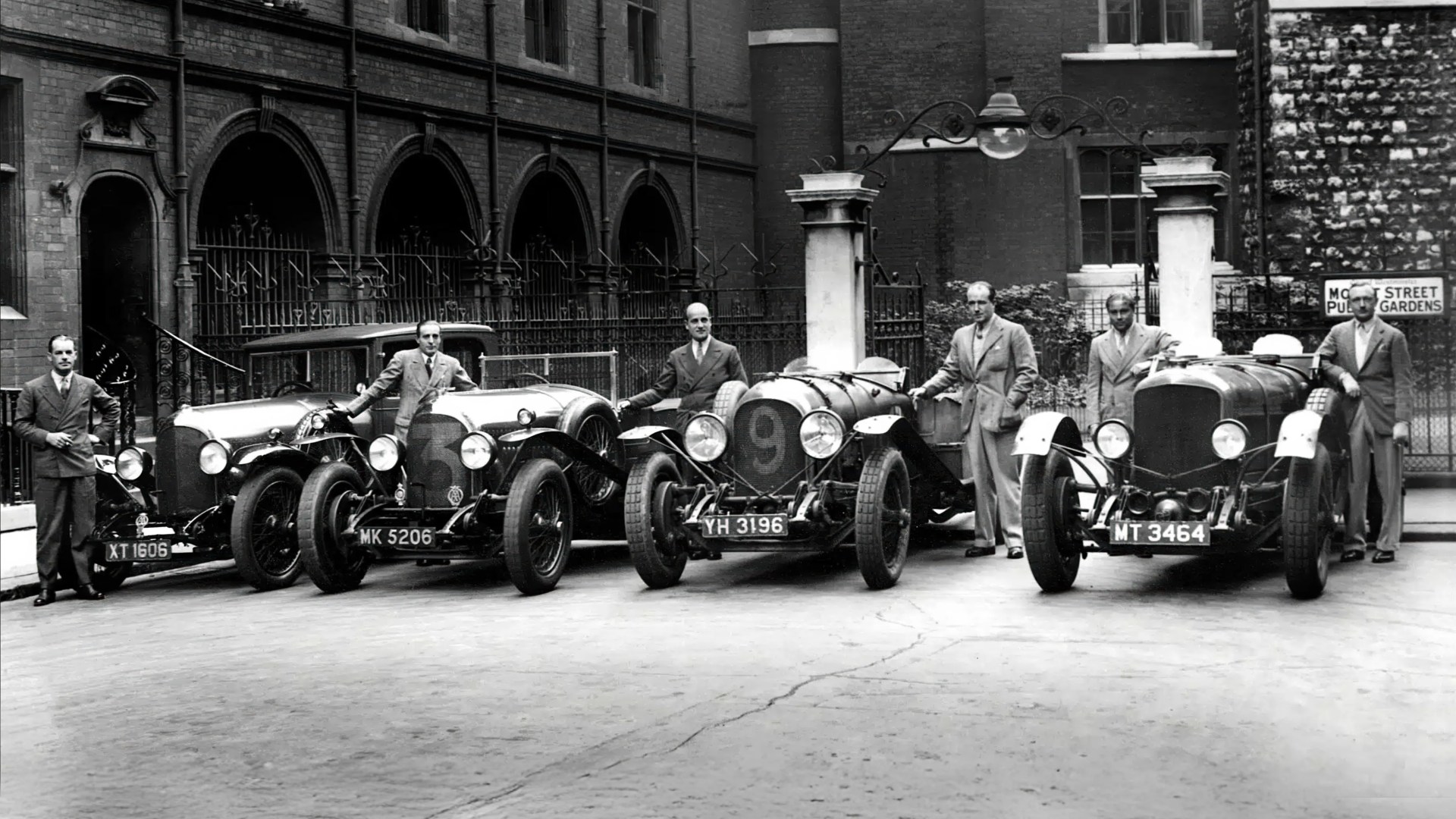 Six men in suits standing beside four vintage race cars with visible license plates in front of a brick building.