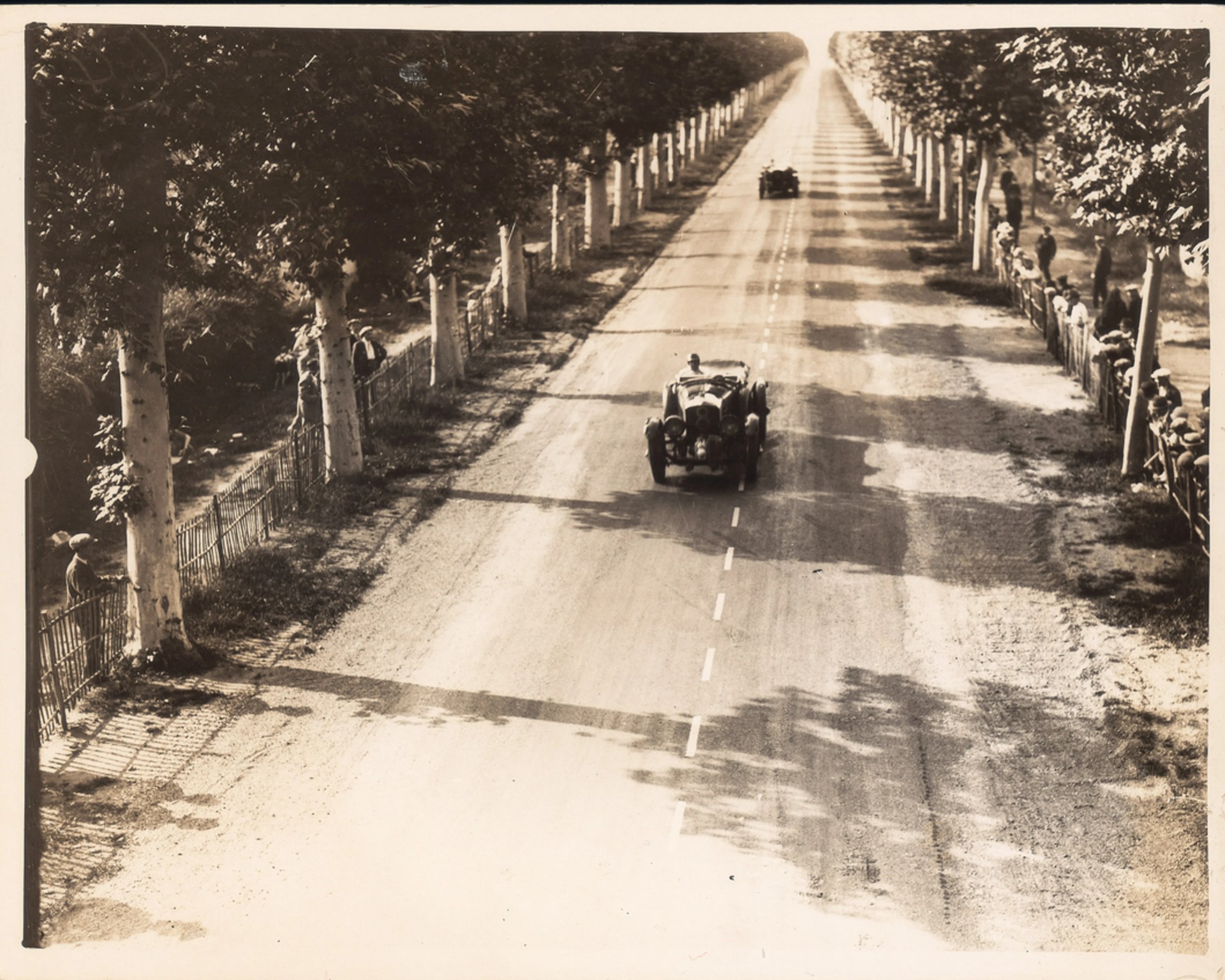 Vintage black-and-white photo of two early 20th-century race cars driving on a tree-lined dirt road with spectators behind fences on both sides.