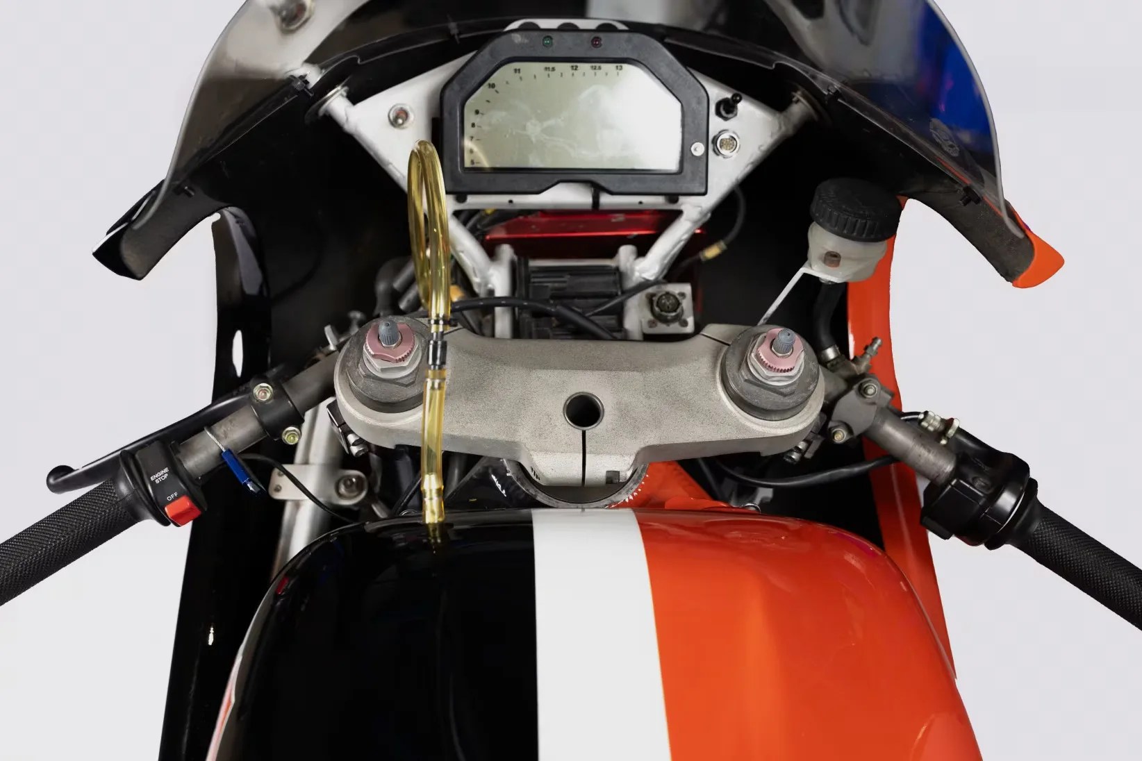 Close-up of a motorcycle dashboard with a digital speedometer, black and orange fuel tank, and handlebar controls.