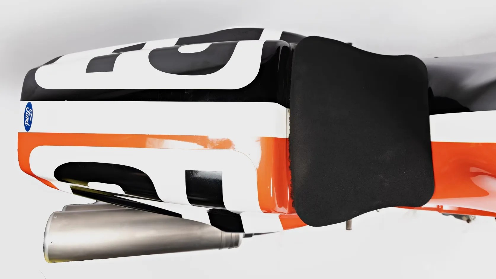 Close-up of a black, white, and orange motorcycle seat with a black foam pad and a silver exhaust pipe underneath.