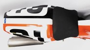 Close-up of a black, white, and orange motorcycle seat with a black foam pad and a silver exhaust pipe underneath.