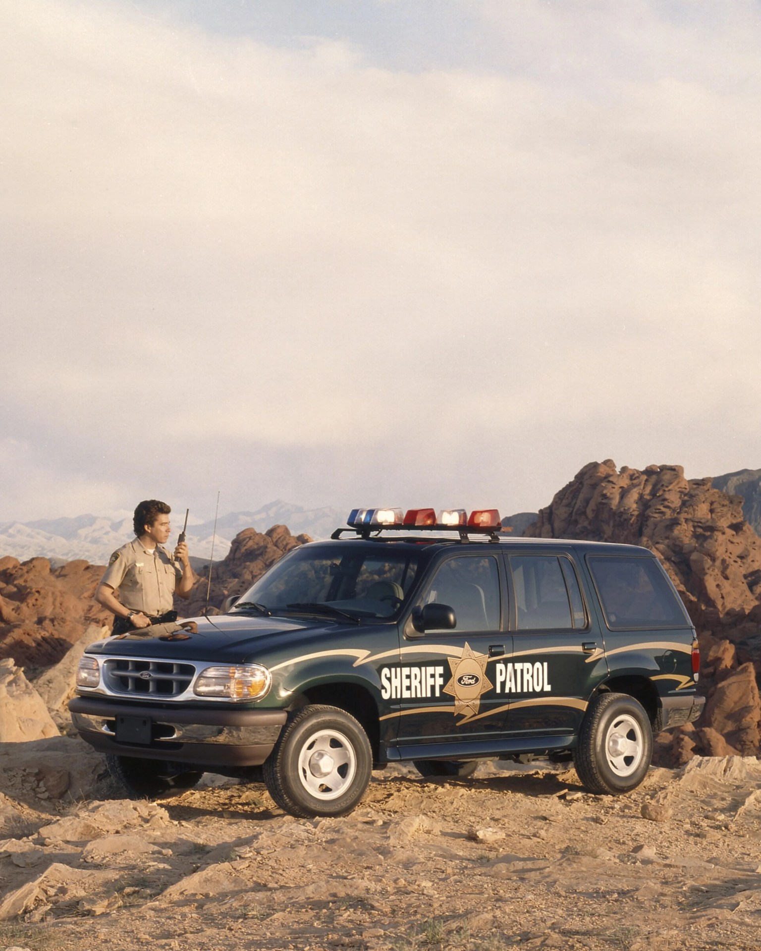 Dark green Ford sheriff patrol SUV with red and blue roof lights parked on rocky terrain with a uniformed officer using a radio.