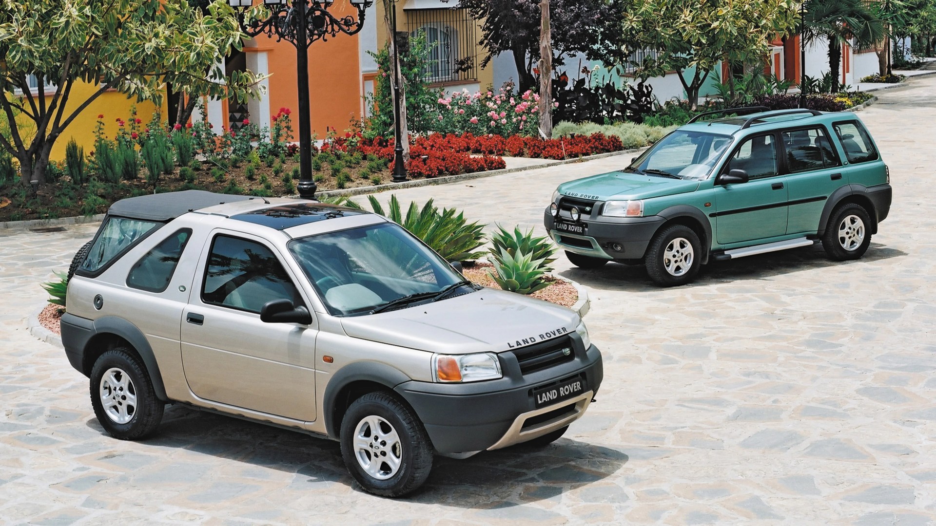 Two Land Rover SUVs, one silver two-door and one green four-door, parked on a stone driveway near colorful houses and plants.