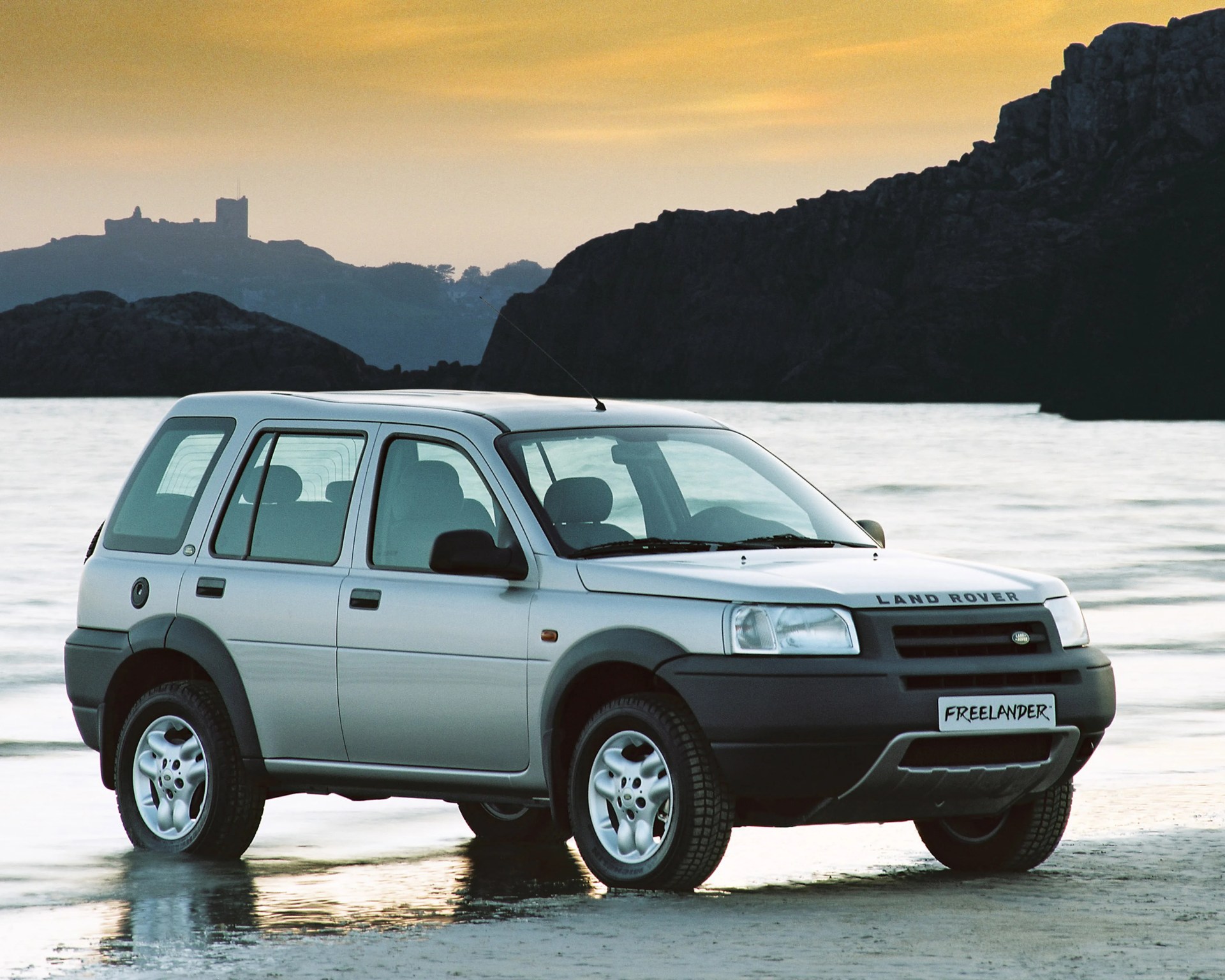 Silver Land Rover Freelander SUV parked on wet sand near the ocean at sunset.