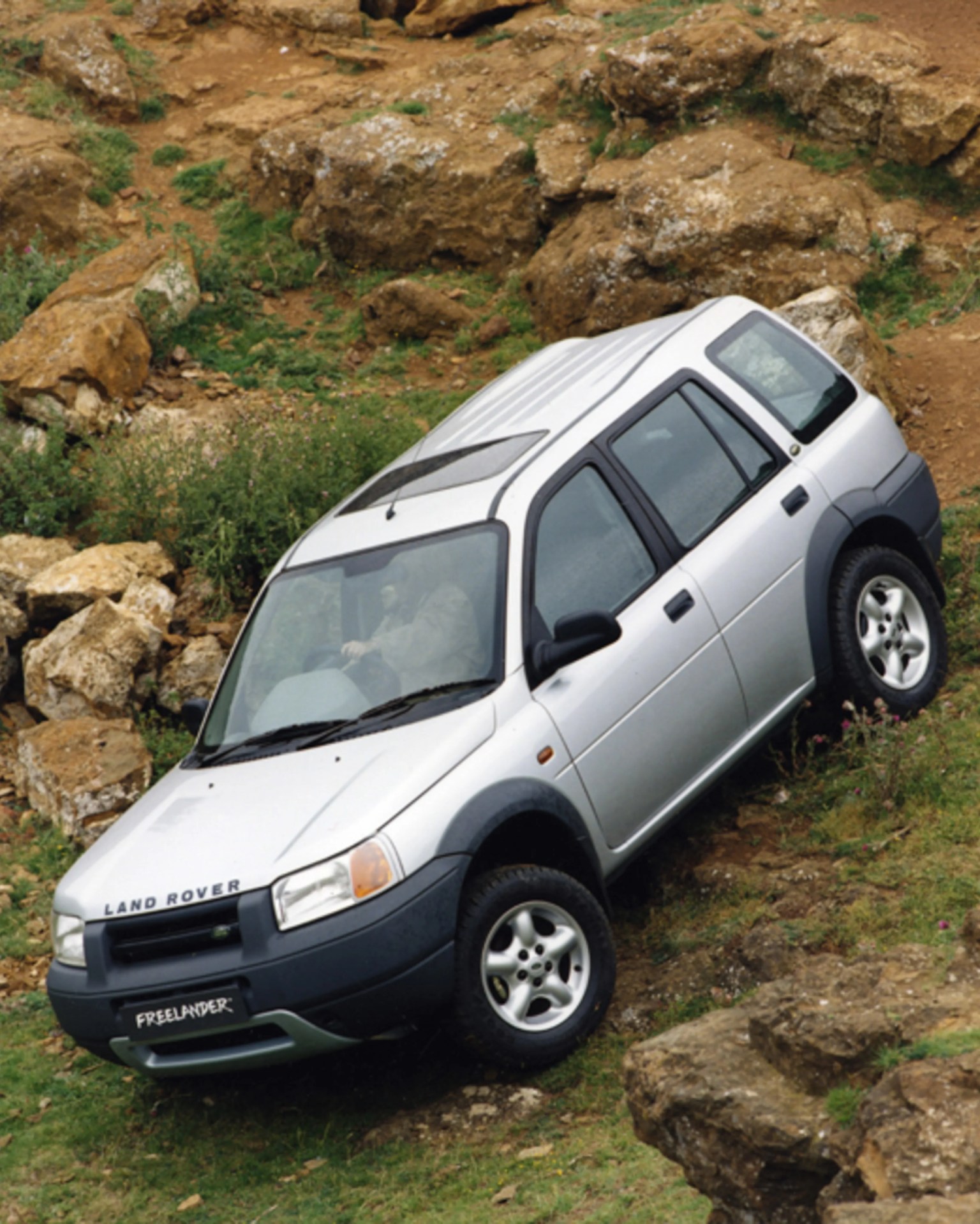 Silver Land Rover Freelander SUV driving on rocky, grassy off-road terrain.