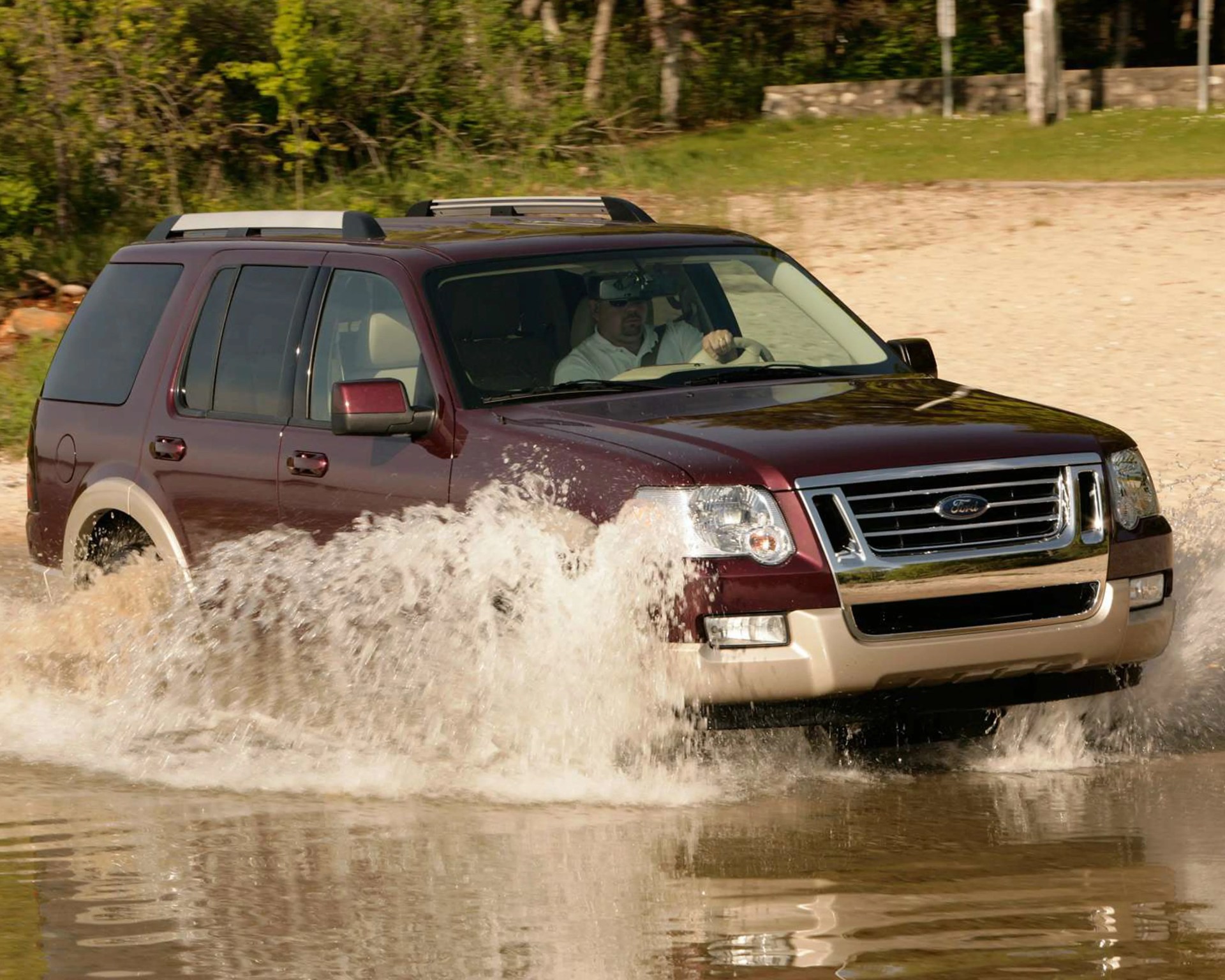 Maroon and beige Ford SUV driving through water splashing on a dirt path with trees in the background.