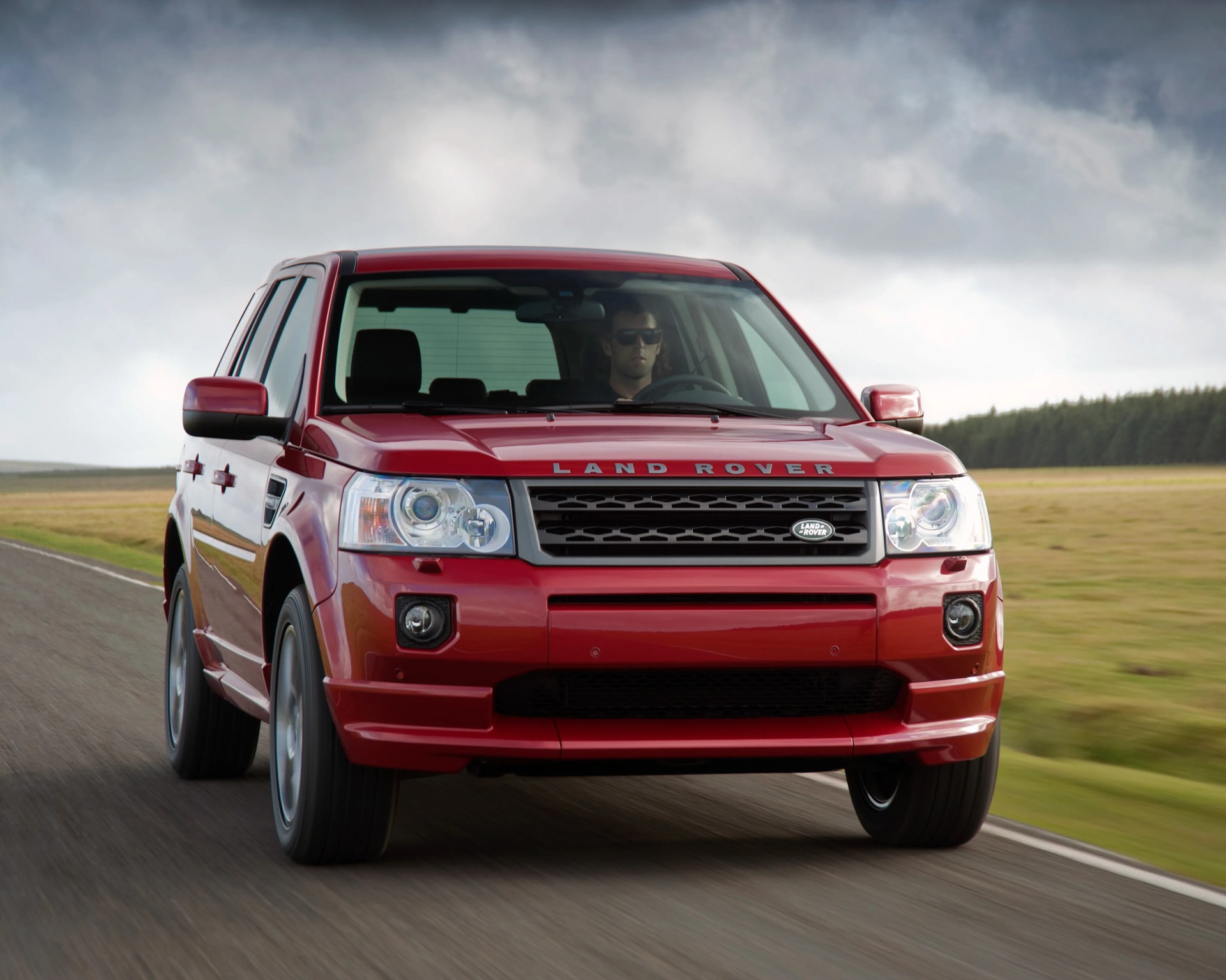 Red Land Rover SUV driving on a rural road under a cloudy sky.