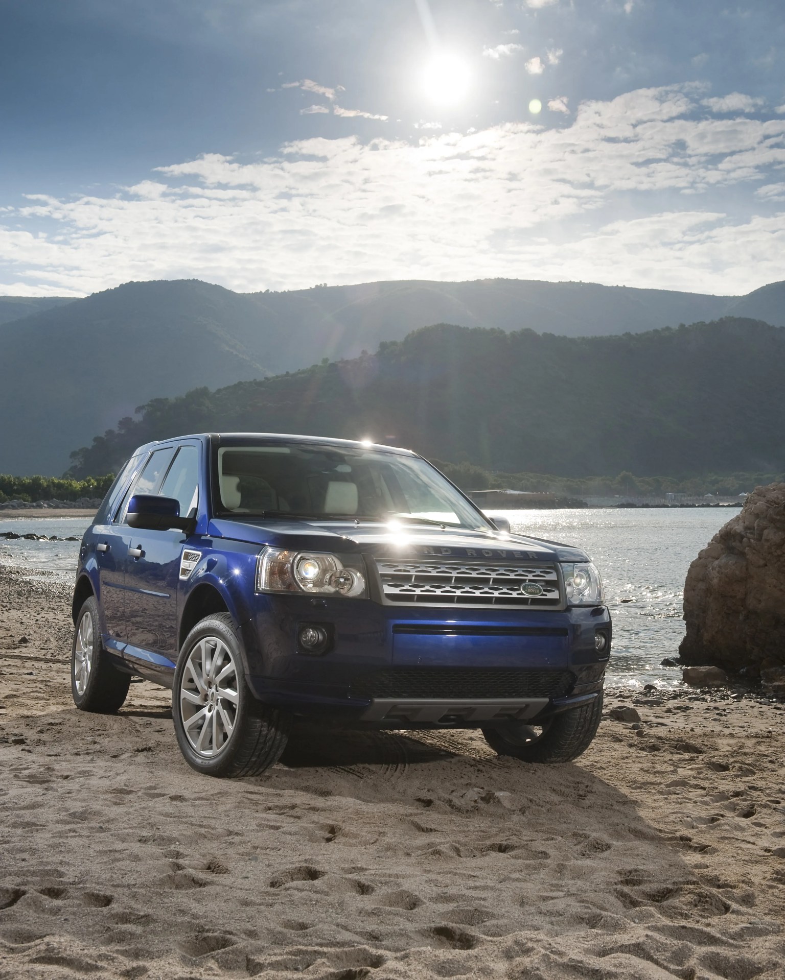 Blue Land Rover SUV parked on sandy beach with mountains and water in the background.