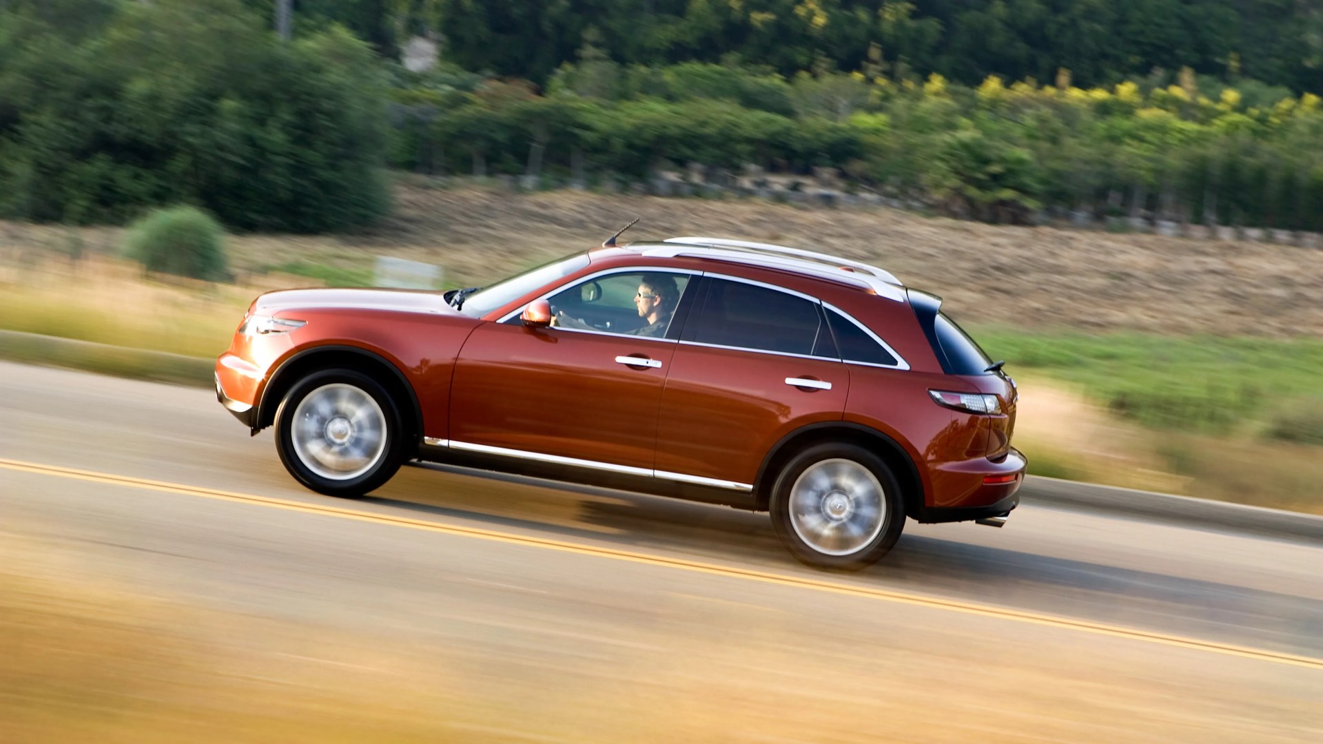 Metallic orange crossover SUV driving on a road with blurred background and spinning wheels.