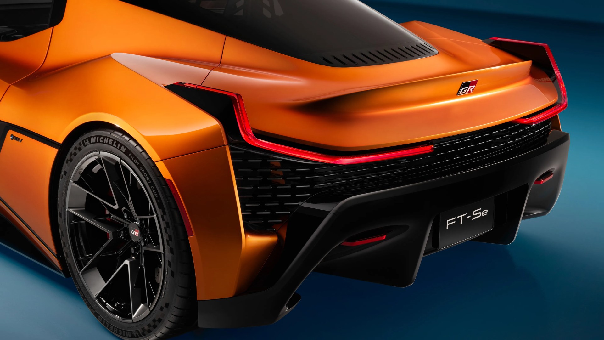 Rear side view of an orange Toyota GR FT-Se sports car with black diffuser and Michelin tires.